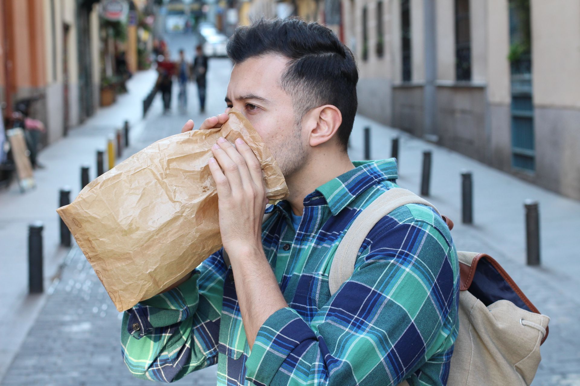 Man breathing into a brown paper bag on a city street, appearing anxious. Man breathing into a brown paper bag on a city street, appearing anxious.