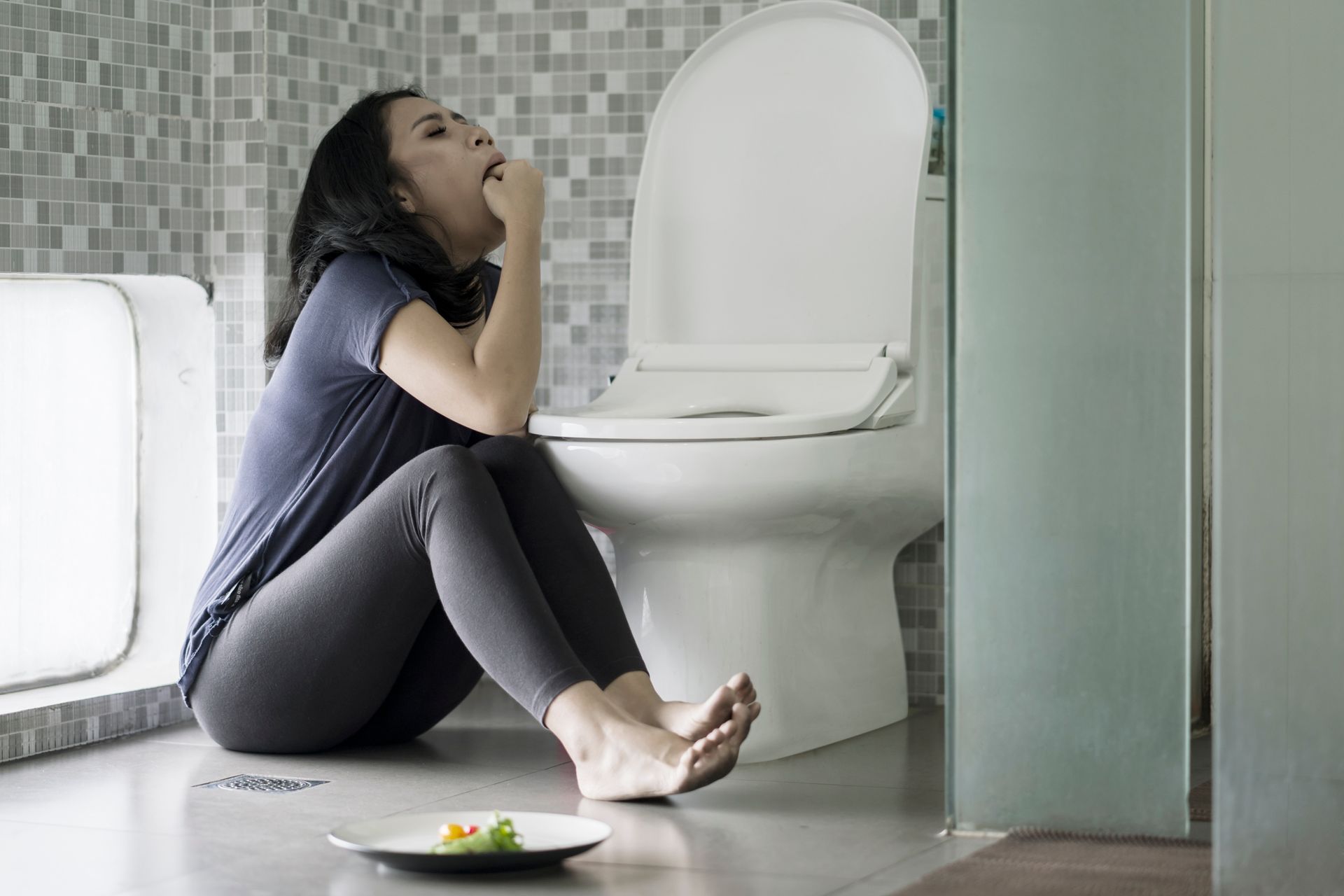 Woman sitting on bathroom floor, leaning toward toilet, looking distressed. Plate of food nearby.
