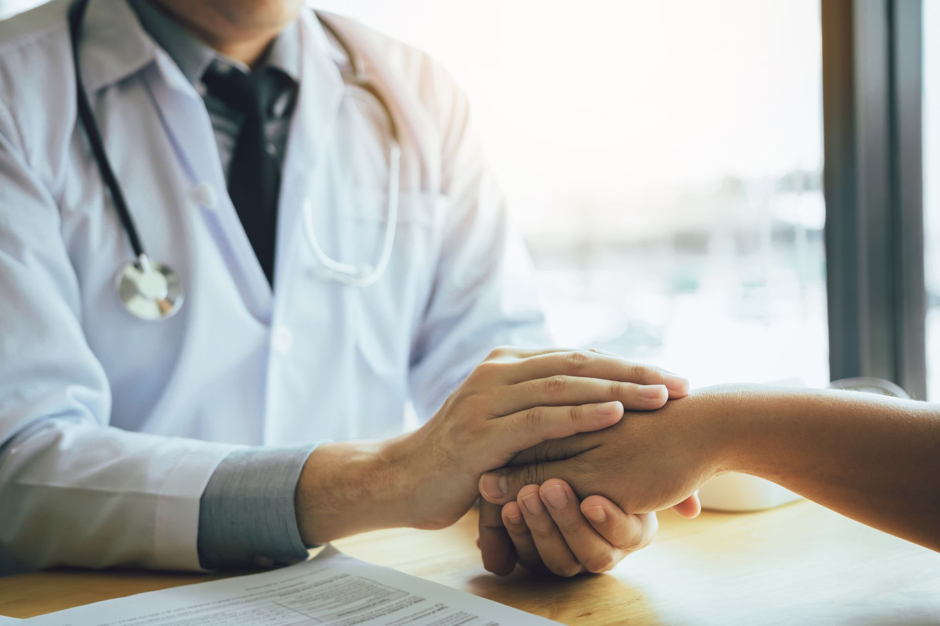 Doctor in white coat holds patient's hand for comfort, in a sunlit room.