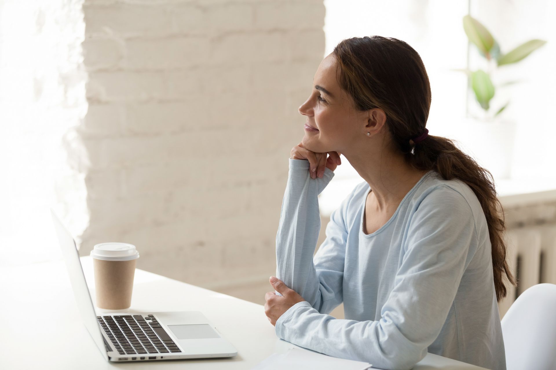 Woman at desk, looking up pensively. Laptop and coffee cup present. Light blue top, white room.