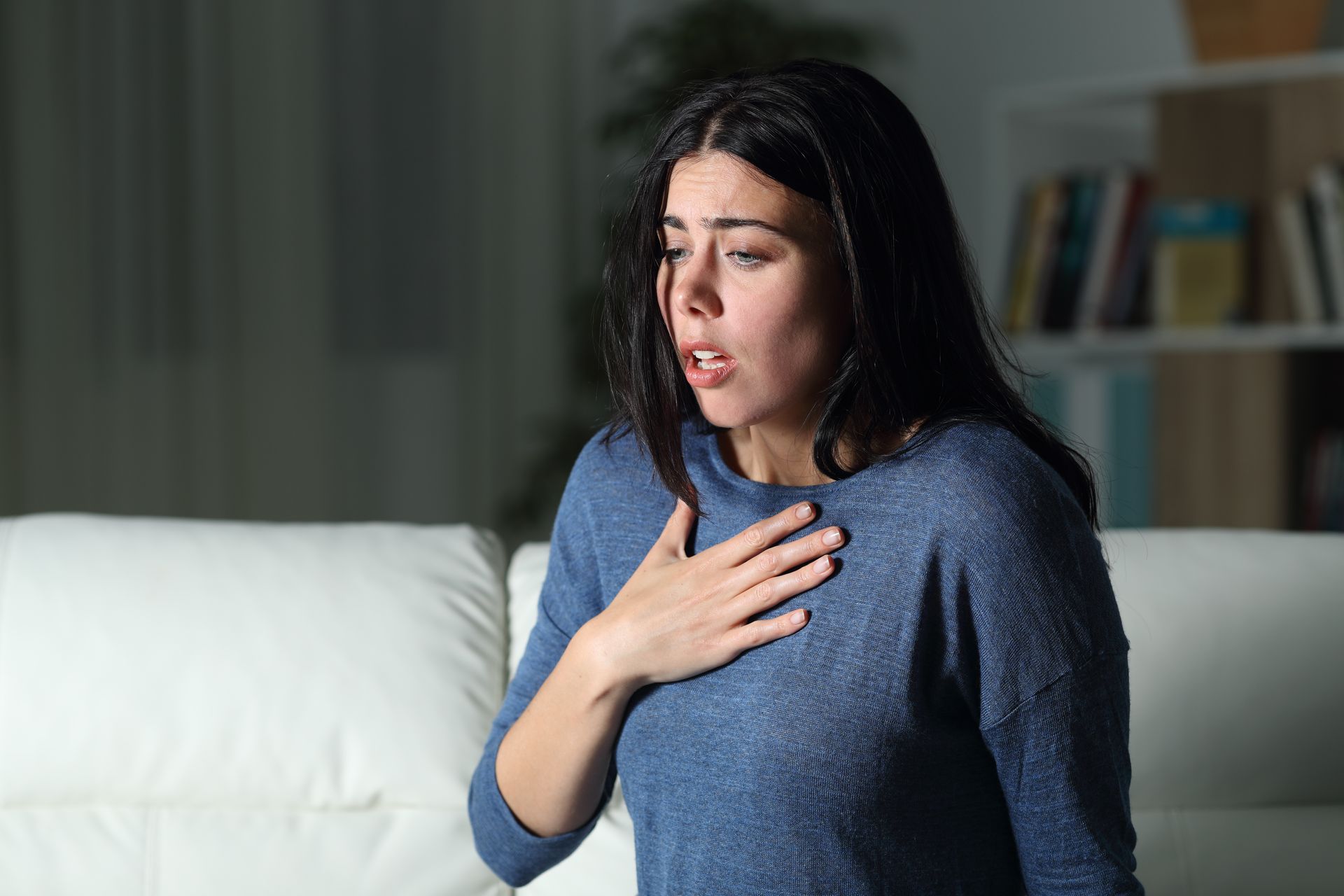 Woman on a couch, clutching chest, mouth open, appearing to struggle to breathe, in a dimly lit room.