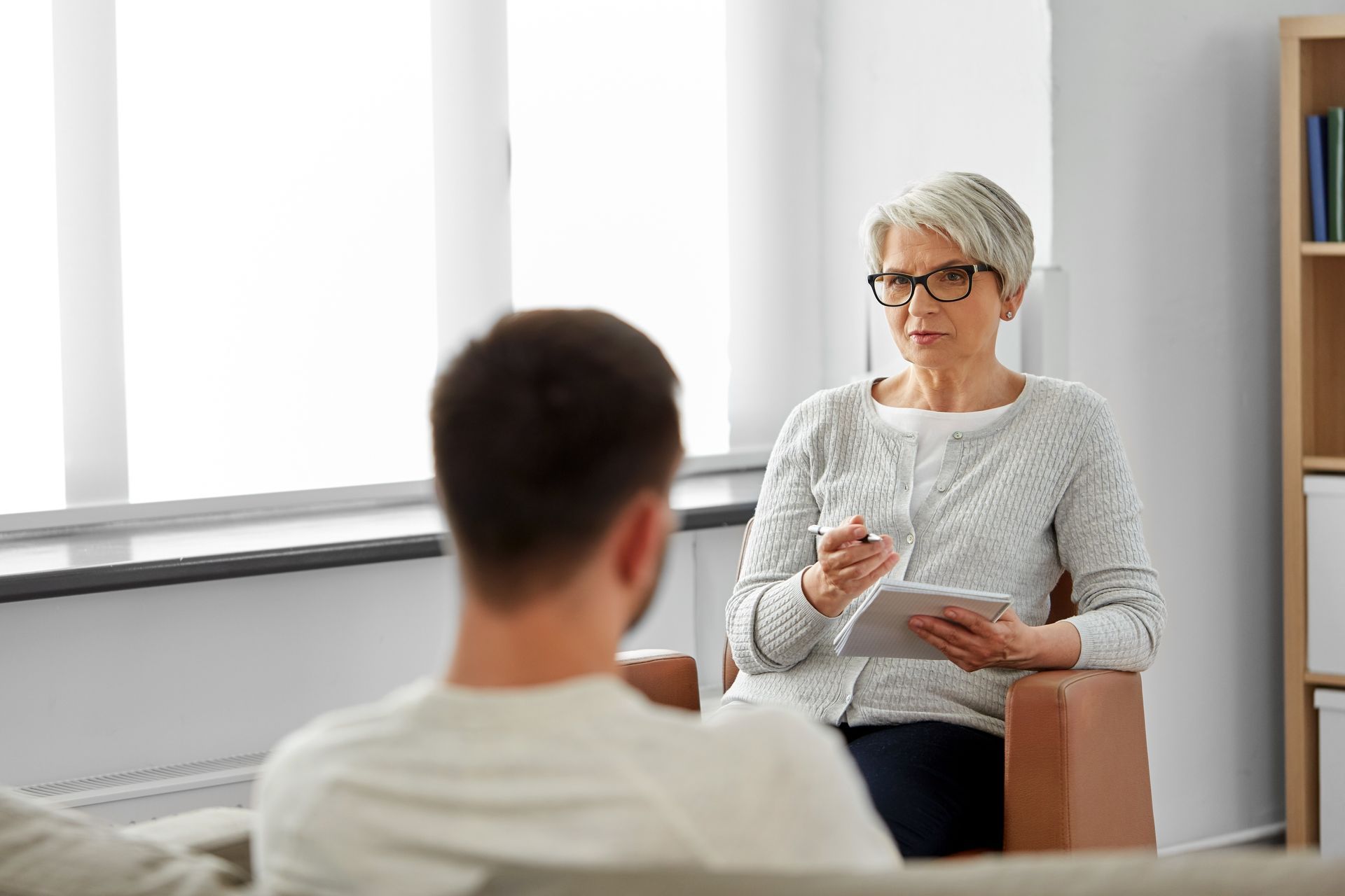 Therapist with glasses holds a tablet, speaking to a person, likely a patient, in an office setting.