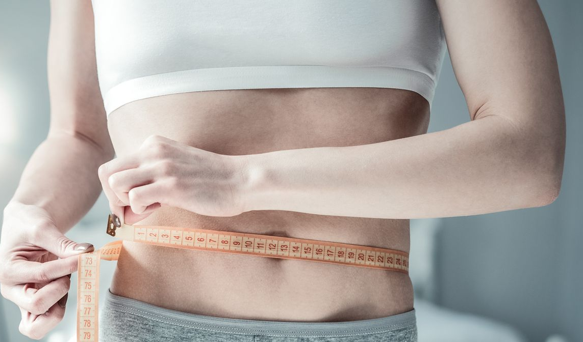 Person in a white crop top and gray shorts measuring waist with a yellow tape measure.