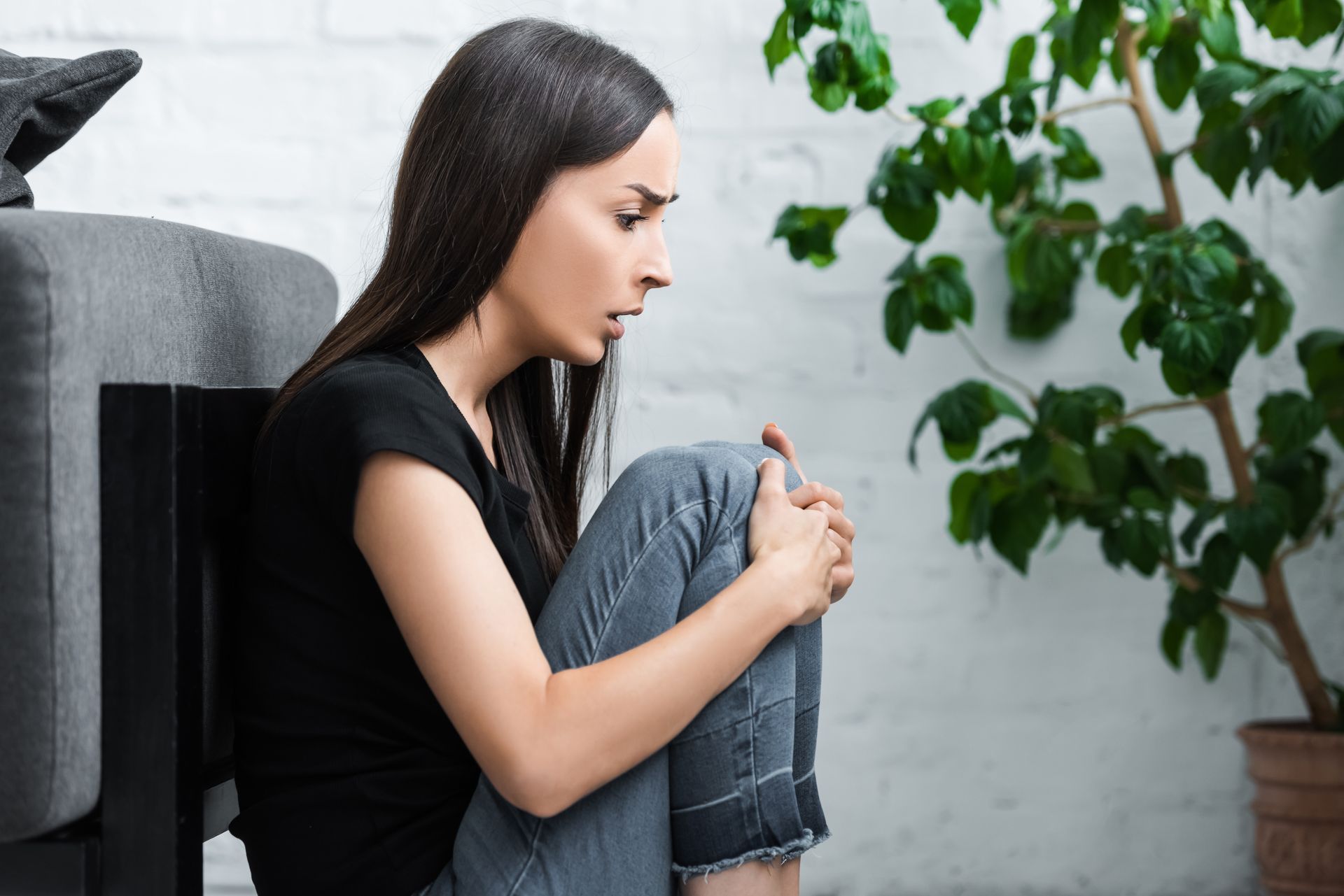 Woman with knees drawn up, sitting on floor, looking distressed. Near a houseplant.