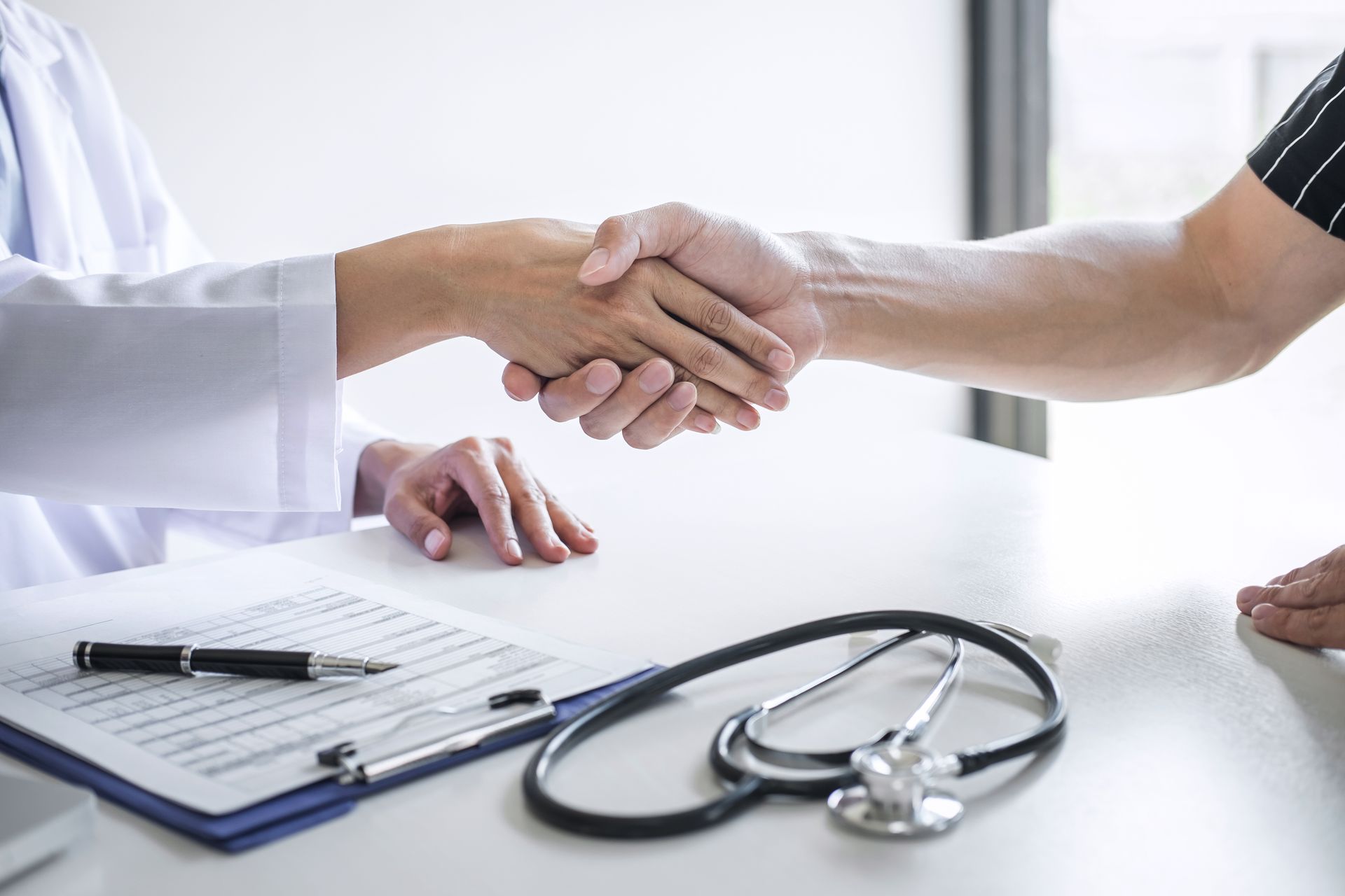Doctor and patient shaking hands at a desk with medical chart and stethoscope.