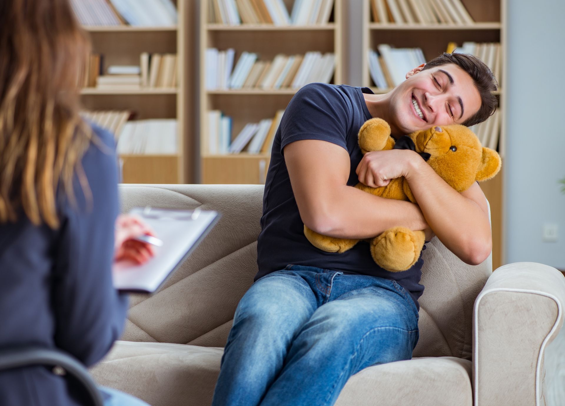 Man hugging teddy bear smiles, in therapy session with counselor, bookshelves in background.