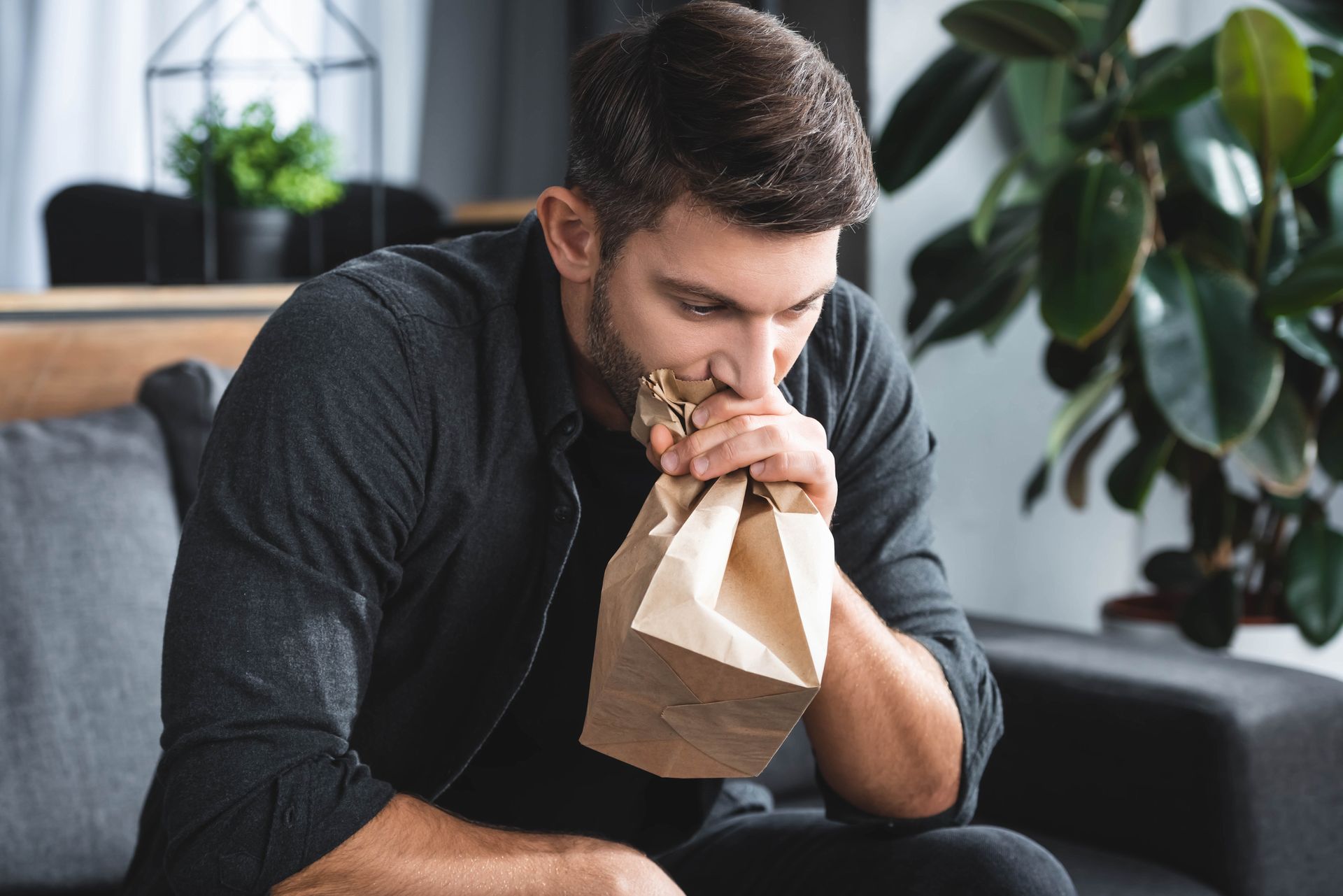 Man on couch breathing into a paper bag, potentially experiencing anxiety in a living room. Man on couch breathing into a paper bag, potentially experiencing anxiety in a living room.