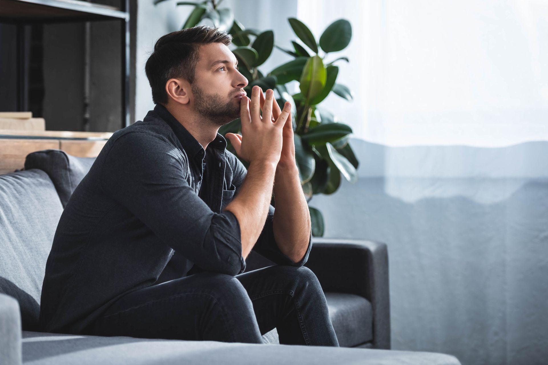 Man sitting on a gray couch, looking up thoughtfully with hands clasped near face, in a room with a window and a plant. Man sitting on a gray couch, looking up thoughtfully with hands clasped near face, in a room with a window and a plant.