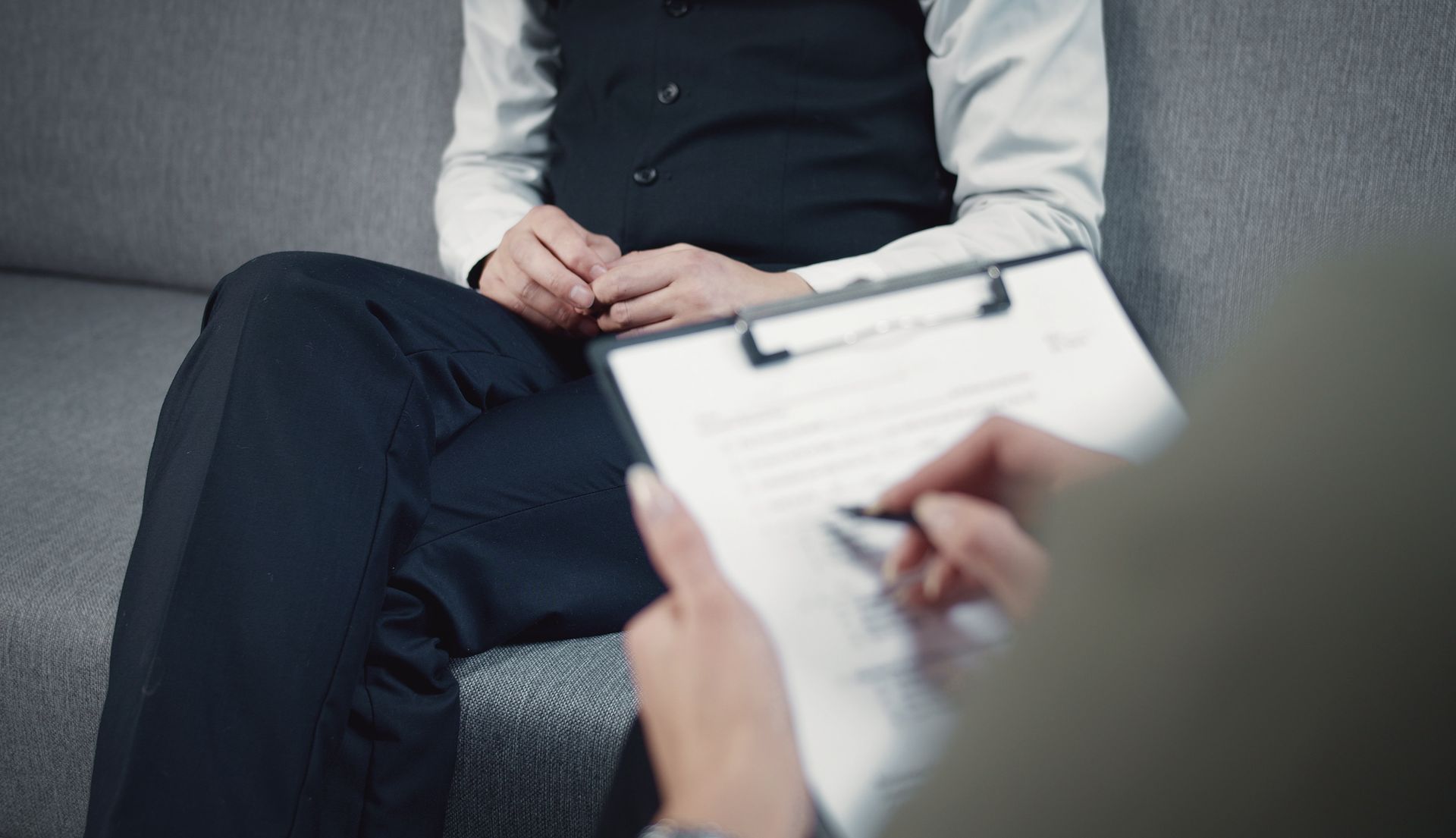 Person in vest sits on couch while another person writes on a clipboard, possibly during a therapy session. Person in vest sits on couch while another person writes on a clipboard, possibly during a therapy session.