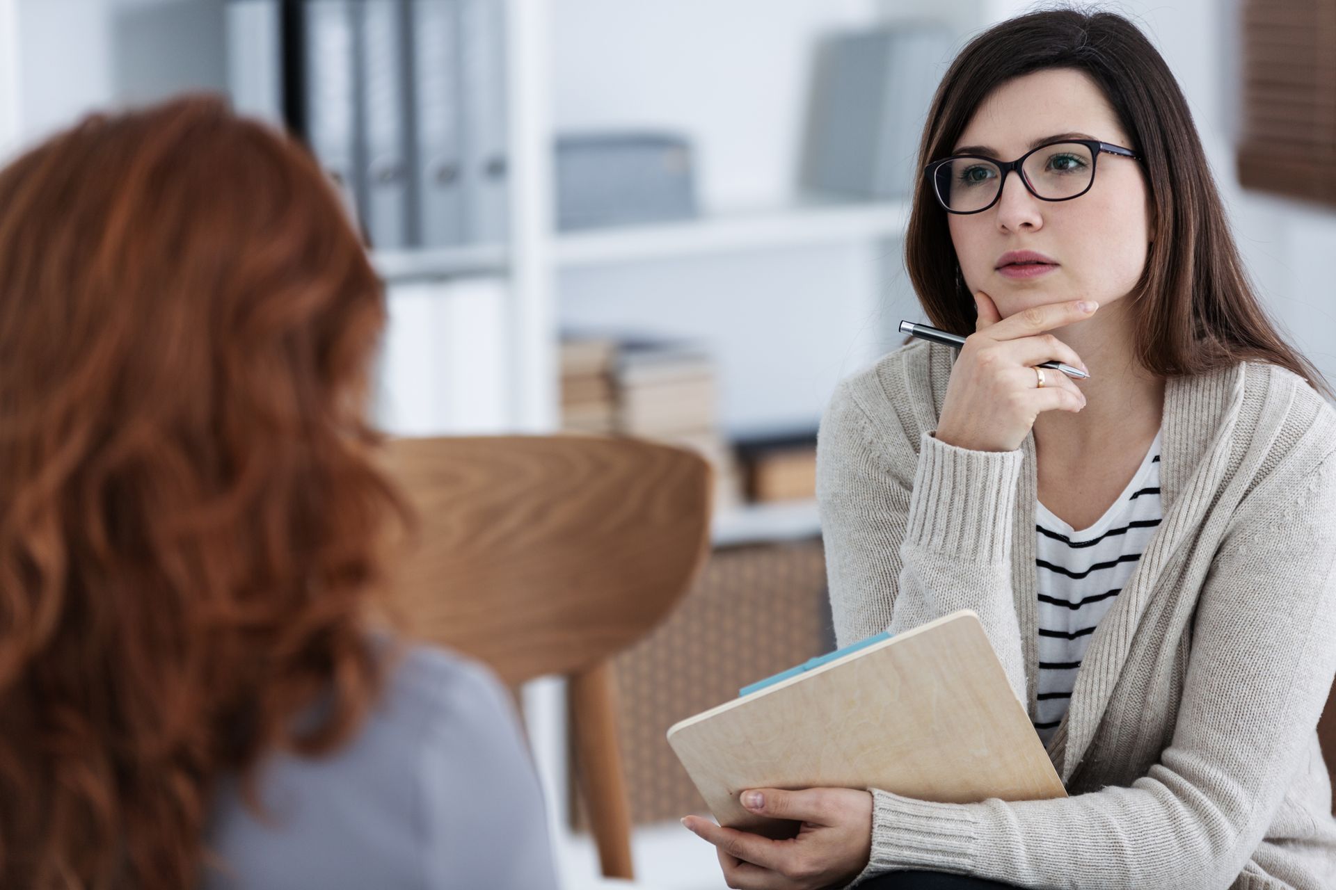 Woman with glasses listens attentively to another woman in a counseling setting.