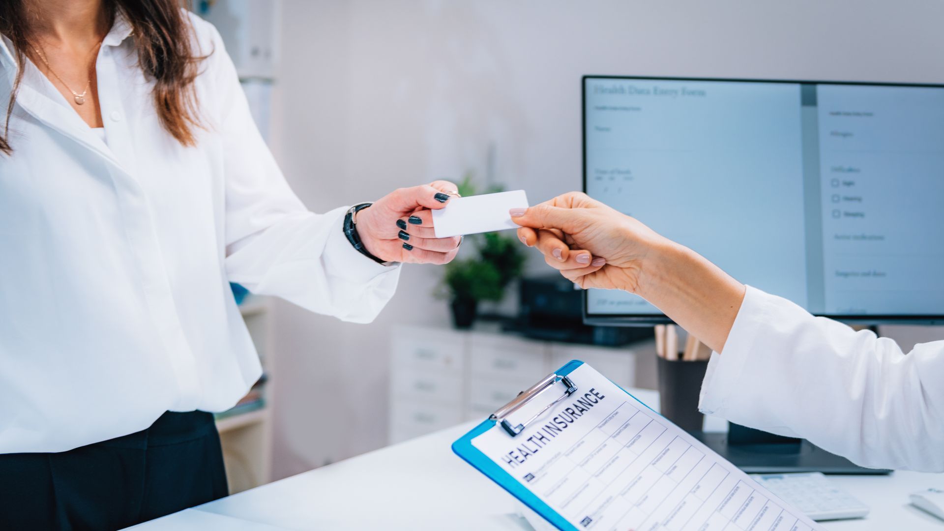 Person in white shirt handing a card to another person behind a desk, healthcare setting. Person in white shirt handing a card to another person behind a desk, healthcare setting.