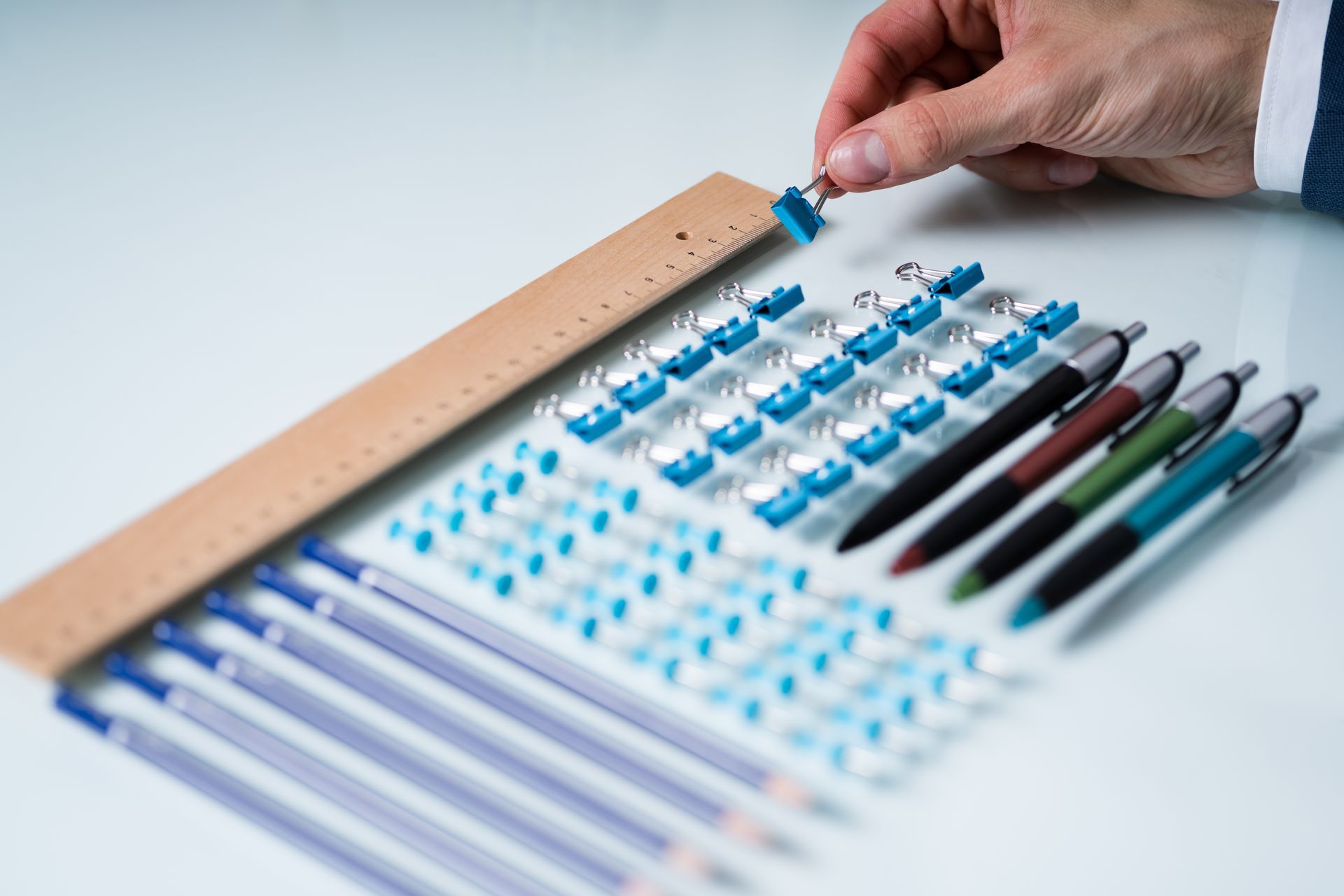 Hand placing a blue push pin next to other push pins, pens, pencils, and a ruler on a white surface.