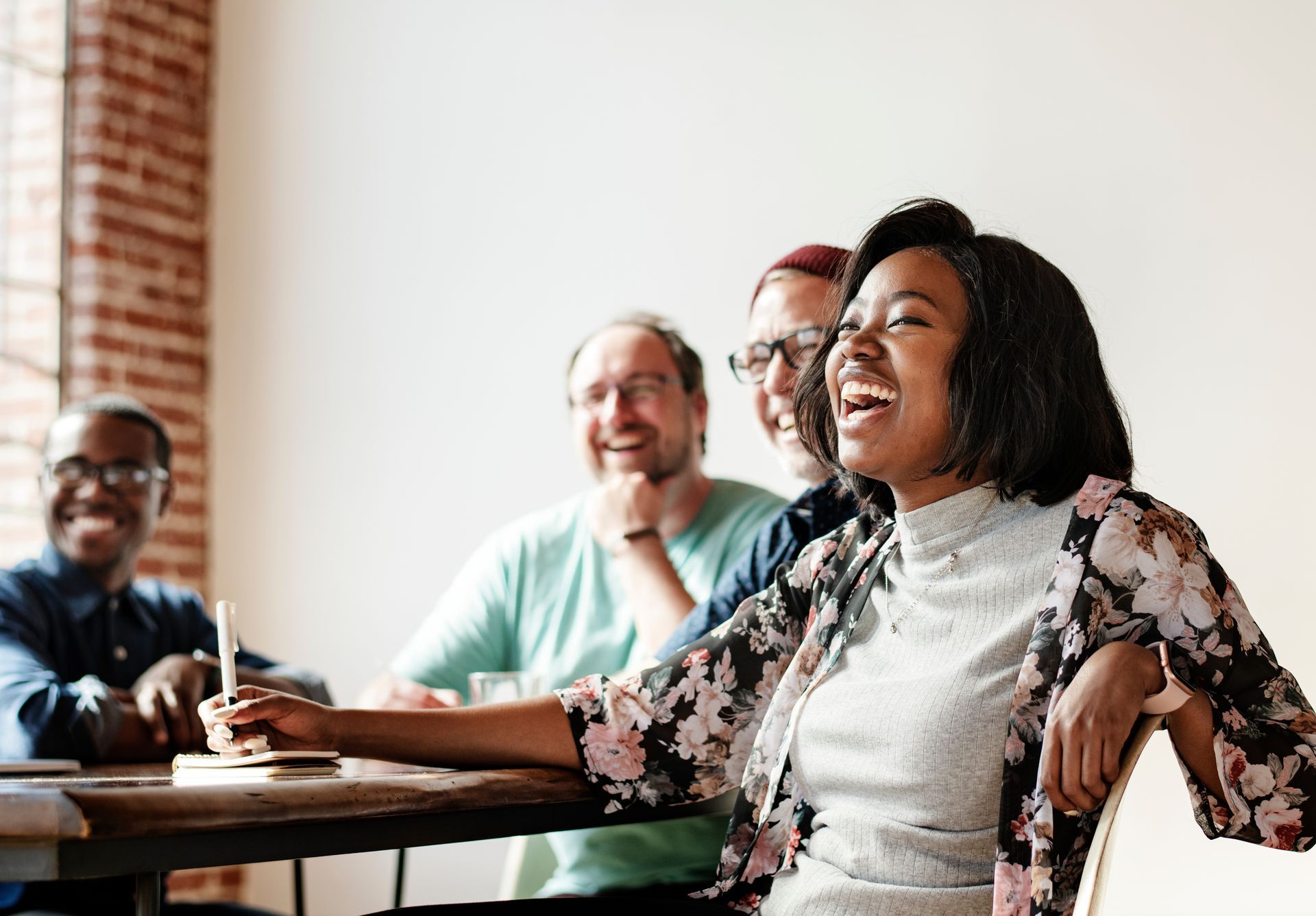 Four people laughing around a table indoors; one woman in a floral jacket is the focal point.