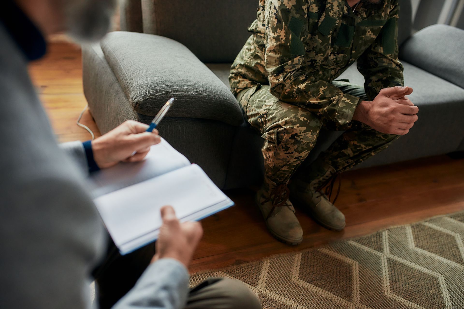 Therapist with notepad and pen; soldier in uniform sits on couch, hands clasped. Counseling session.