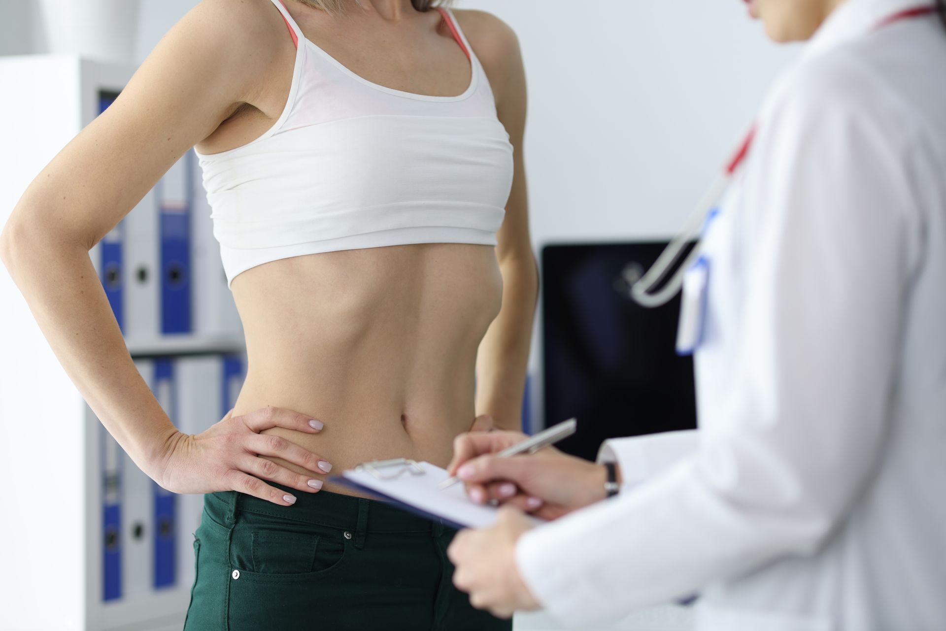 Woman in white top and green pants being examined by a doctor with clipboard in office.