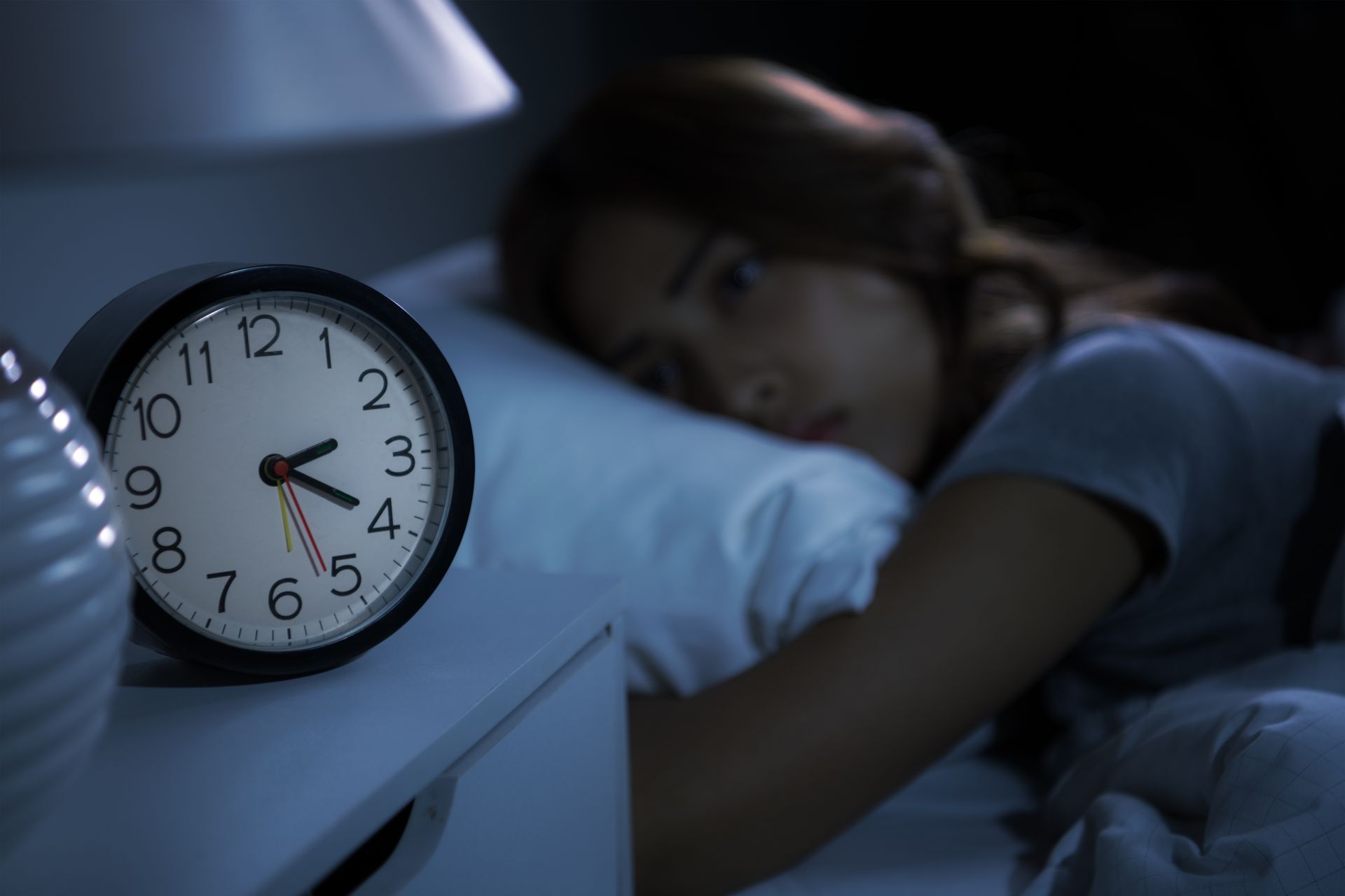 Alarm clock on a nightstand with a woman lying in bed, appearing awake.