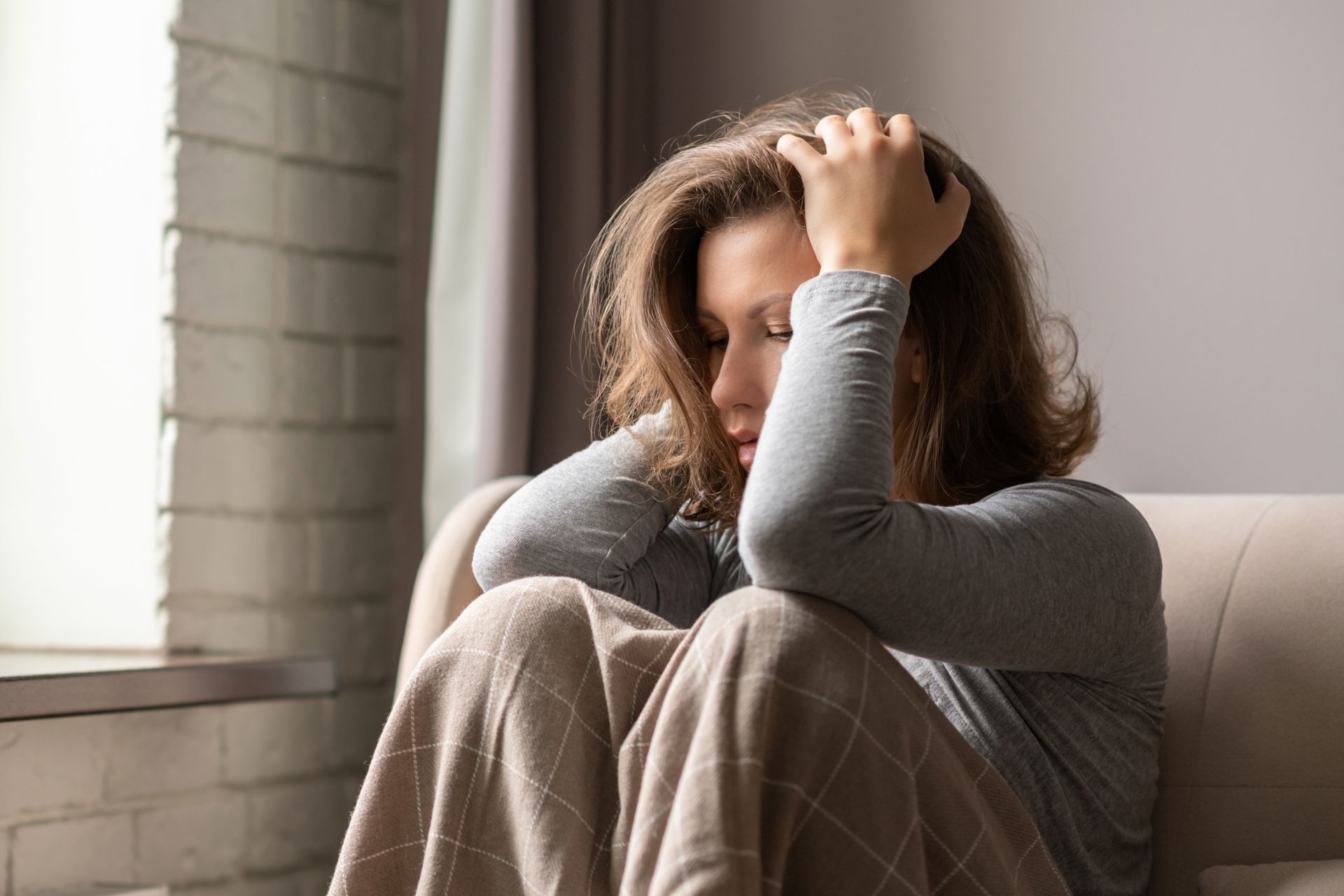 Woman sitting on a couch, holding her head with a look of distress. She is near a window.