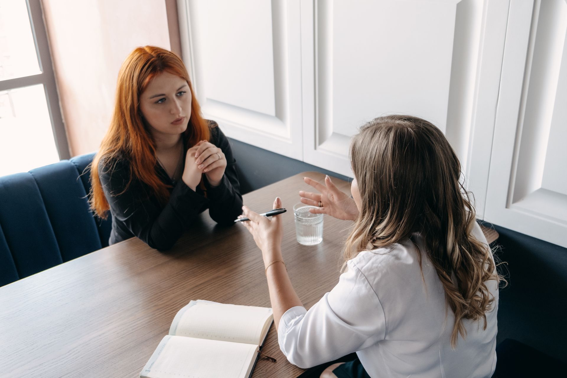 Two women at a table, one gesturing with a pen, the other looking concerned. Indoors, natural light. Two women at a table, one gesturing with a pen, the other looking concerned. Indoors, natural light.