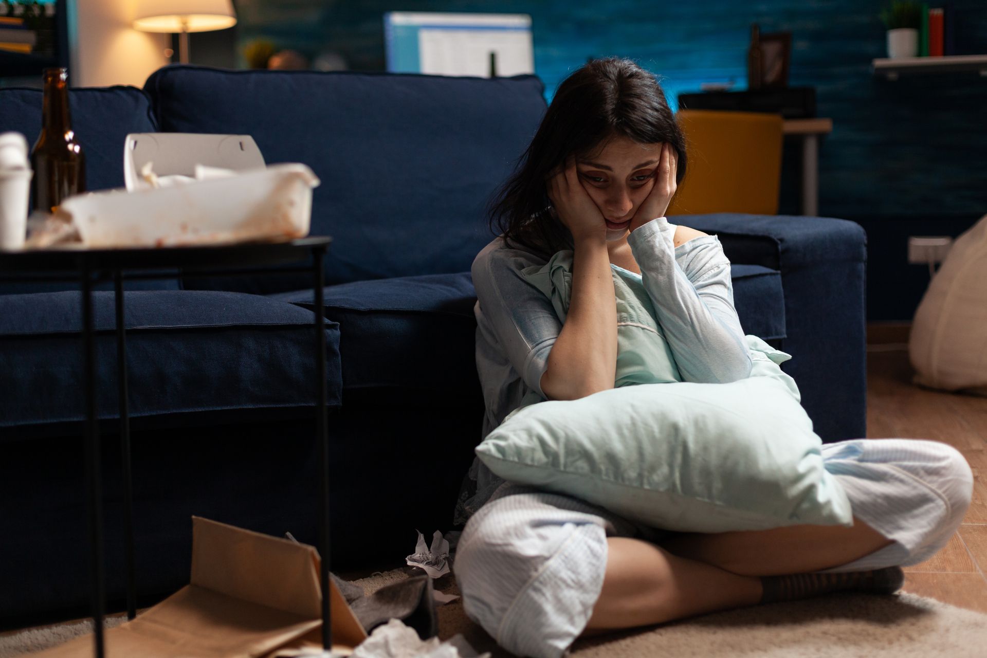Woman sitting on the floor, holding a pillow, looking sad. Surrounded by trash and a couch. Woman sitting on the floor, holding a pillow, looking sad. Surrounded by trash and a couch.