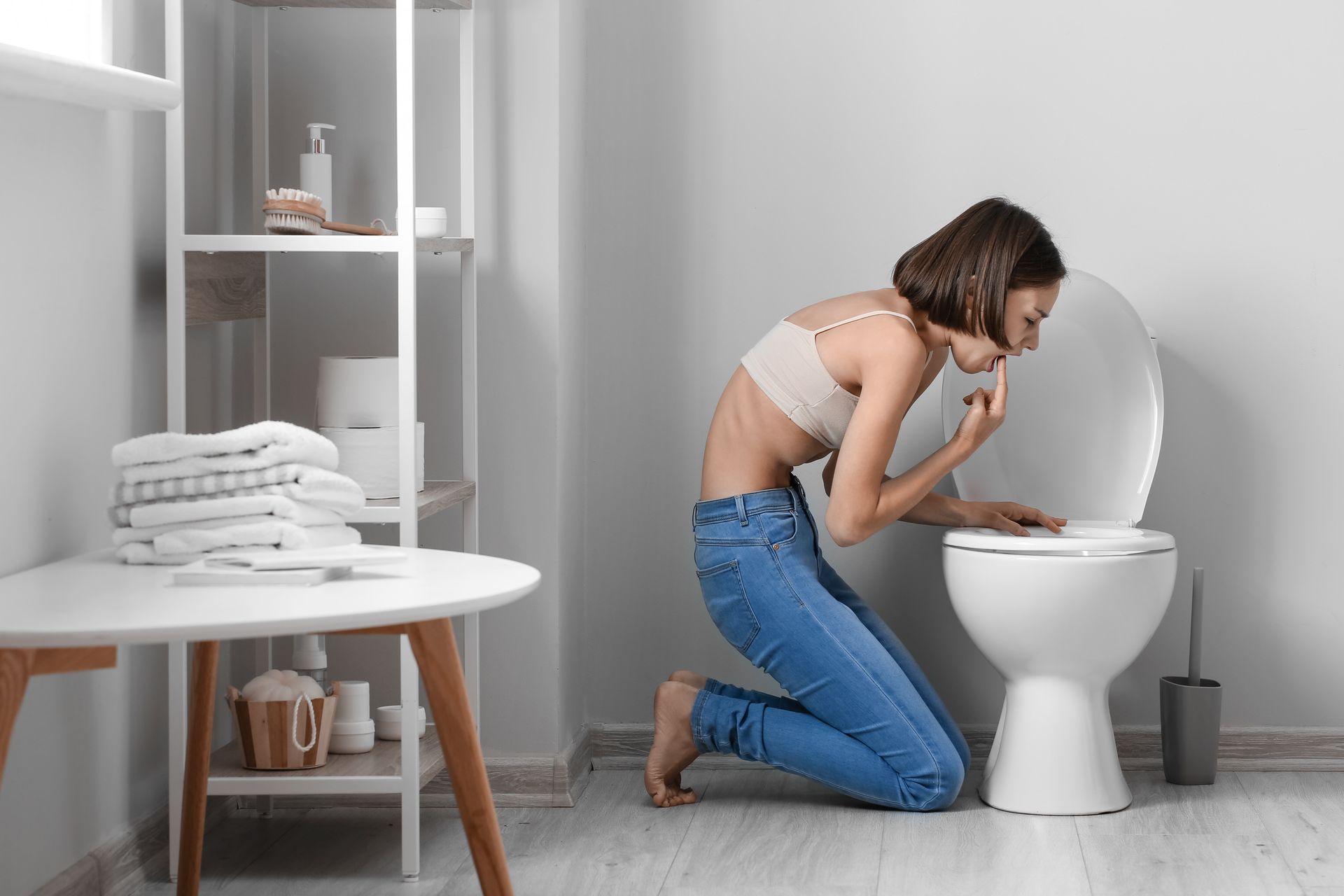 Woman in jeans and bra bent over toilet, looking ill. Interior bathroom with towels and shelves.