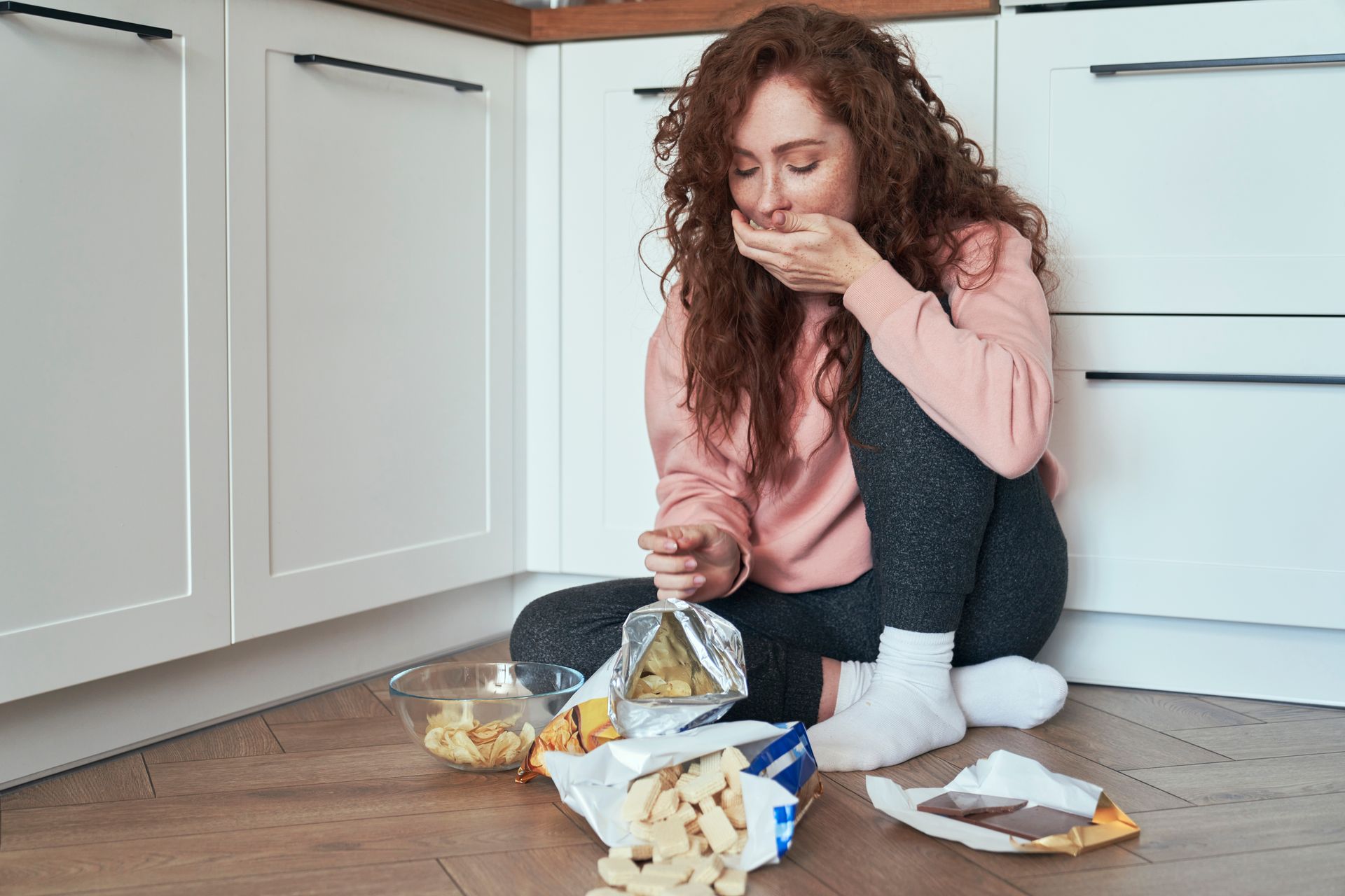 Woman on kitchen floor, eating chips from open bag, looking down. Bowl and other food also on floor.