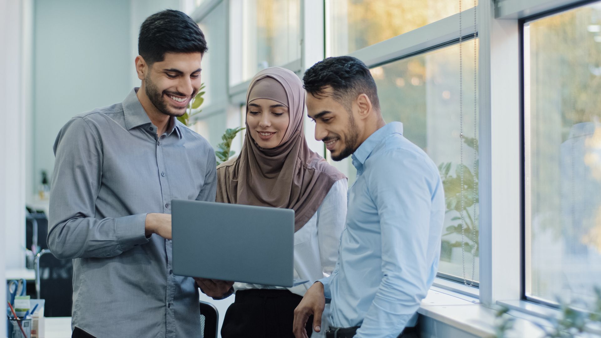 Three people looking at a laptop, smiling.  Near a window, inside.