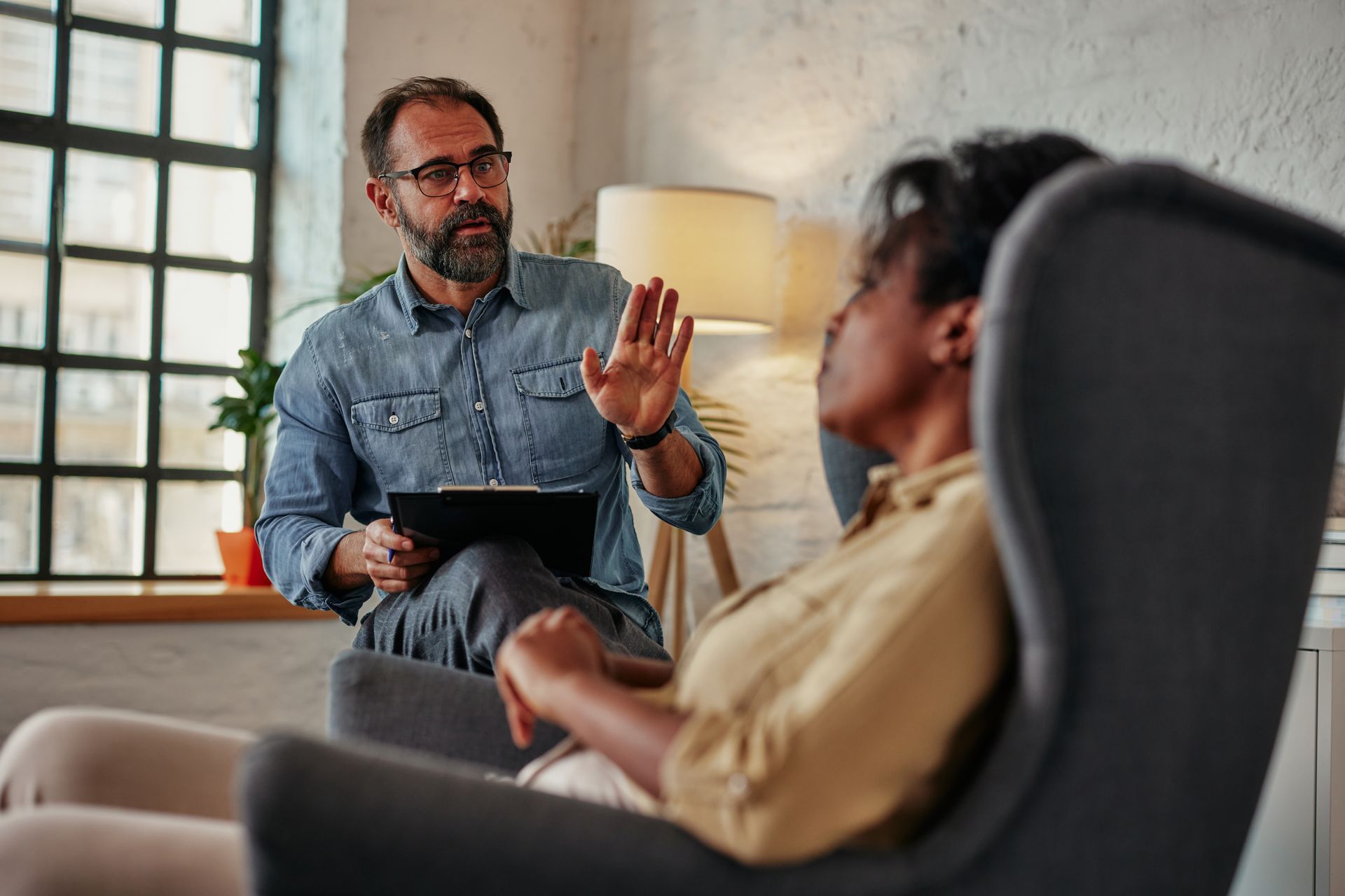 Therapist gesturing while talking to a client in an armchair in a sunlit room.