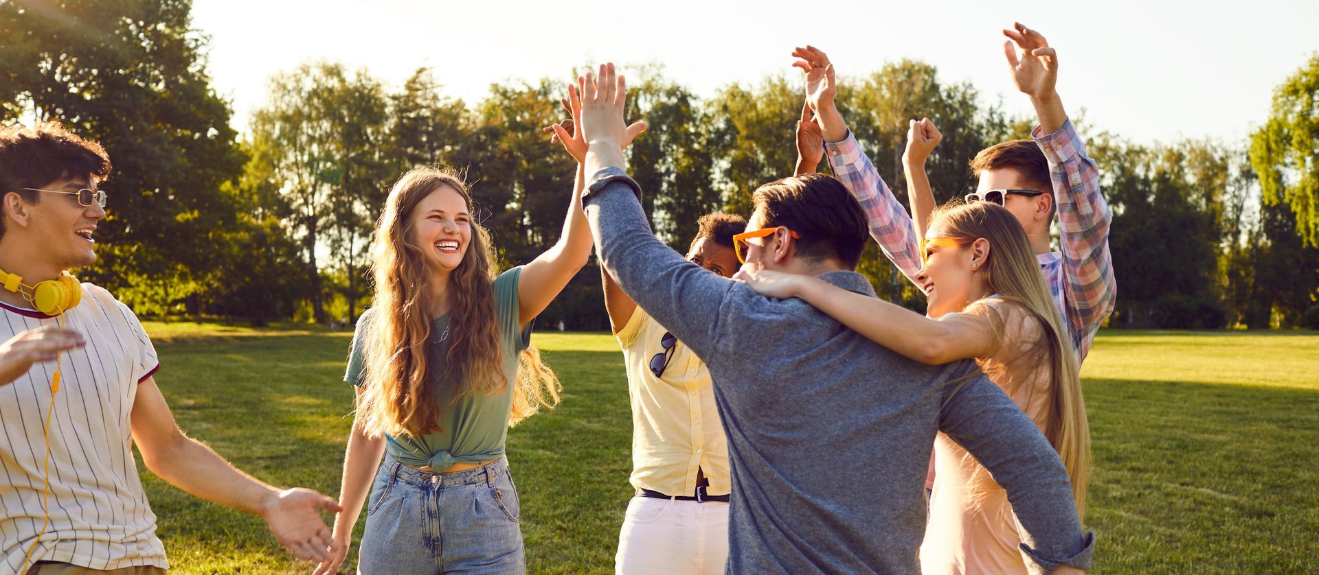 Group of people celebrating in a park, raising their arms and smiling. Sunlight, green grass, and trees.