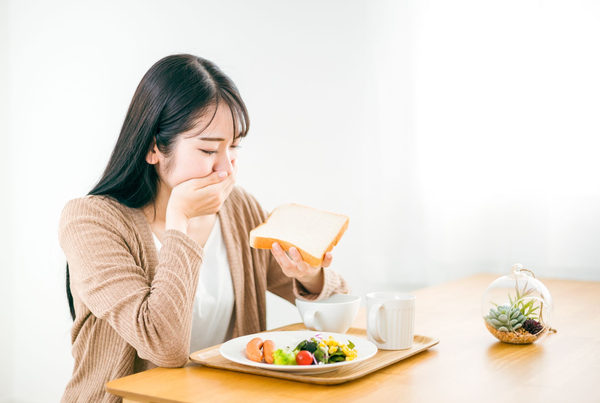 Woman at table with breakfast, covering mouth, holding bread.