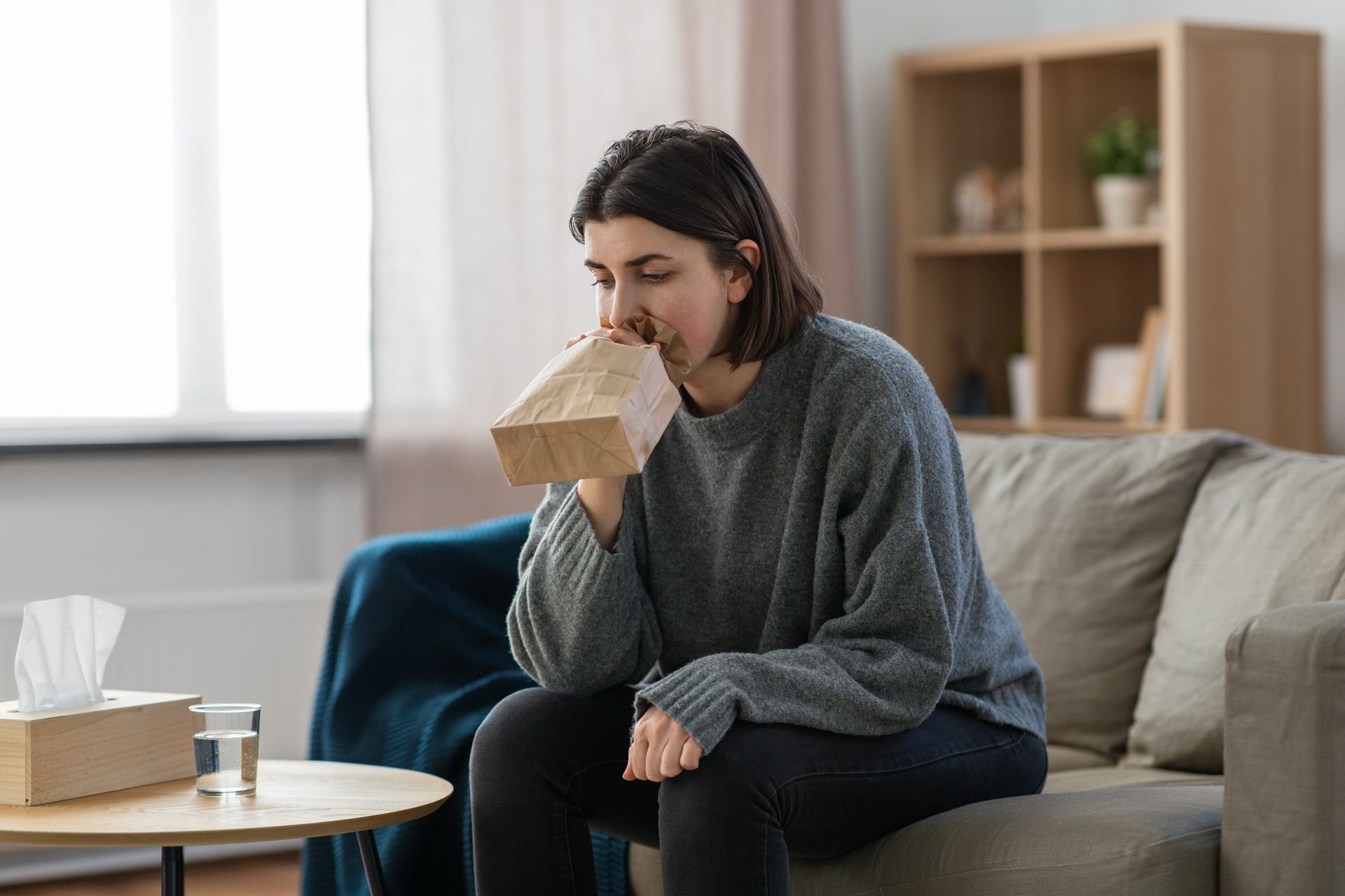 Woman breathing into paper bag on a couch, potentially hyperventilating. Tissue box and water on table nearby.