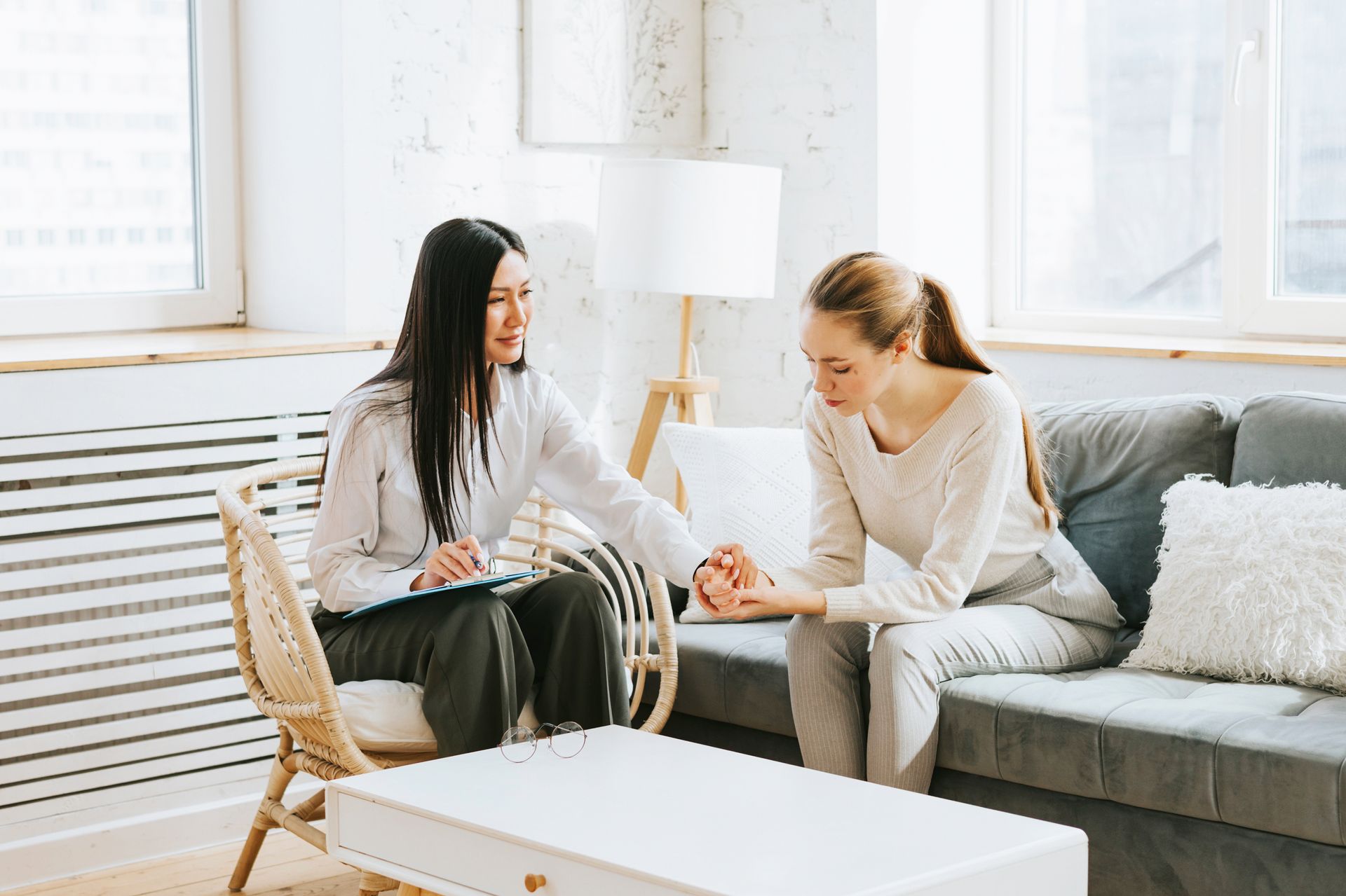 Woman in therapy comforts another woman, holding her hand. They sit in a brightly lit room. Woman in therapy comforts another woman, holding her hand. They sit in a brightly lit room.