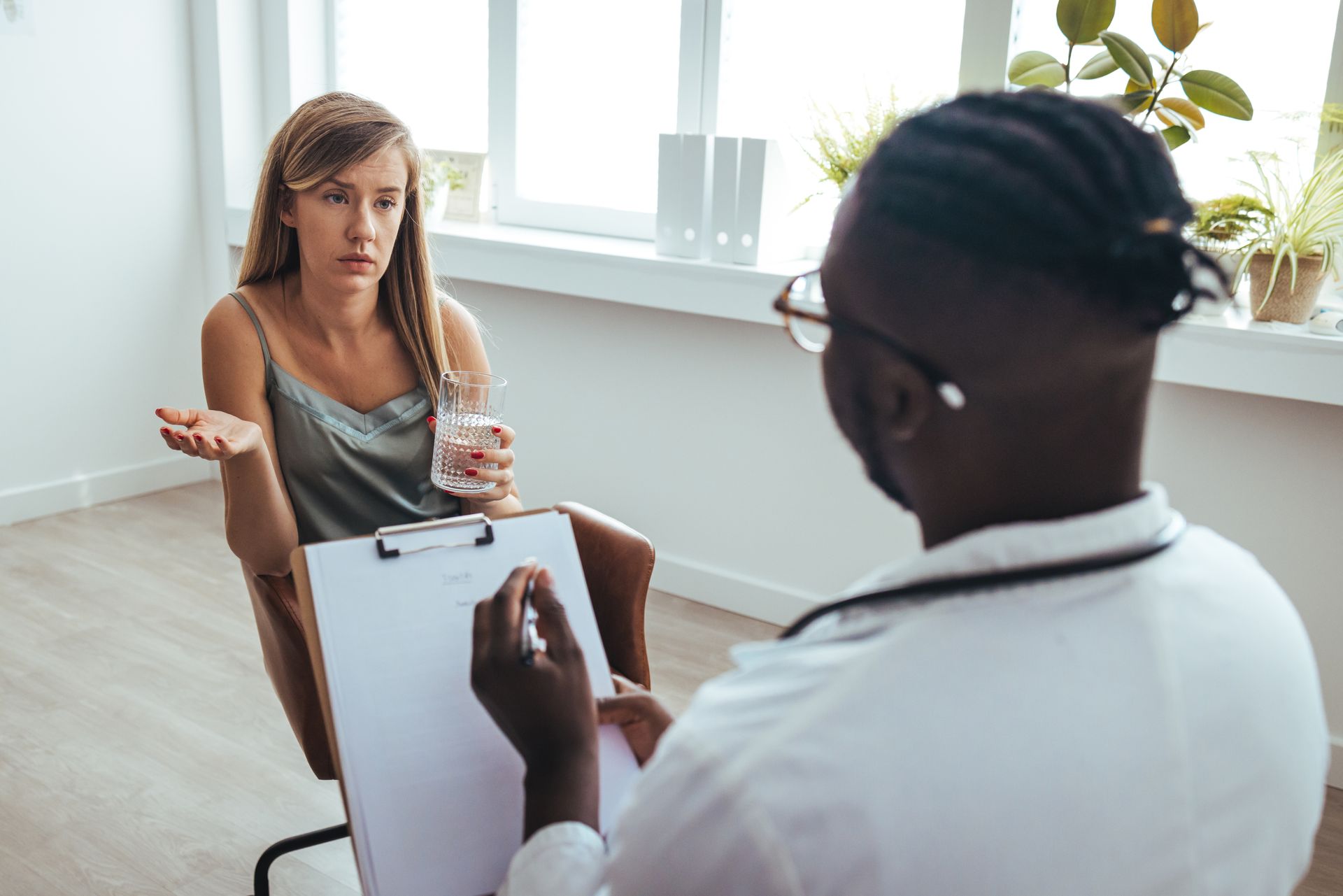 Woman in a therapy session talking to a doctor holding a clipboard; indoor setting.