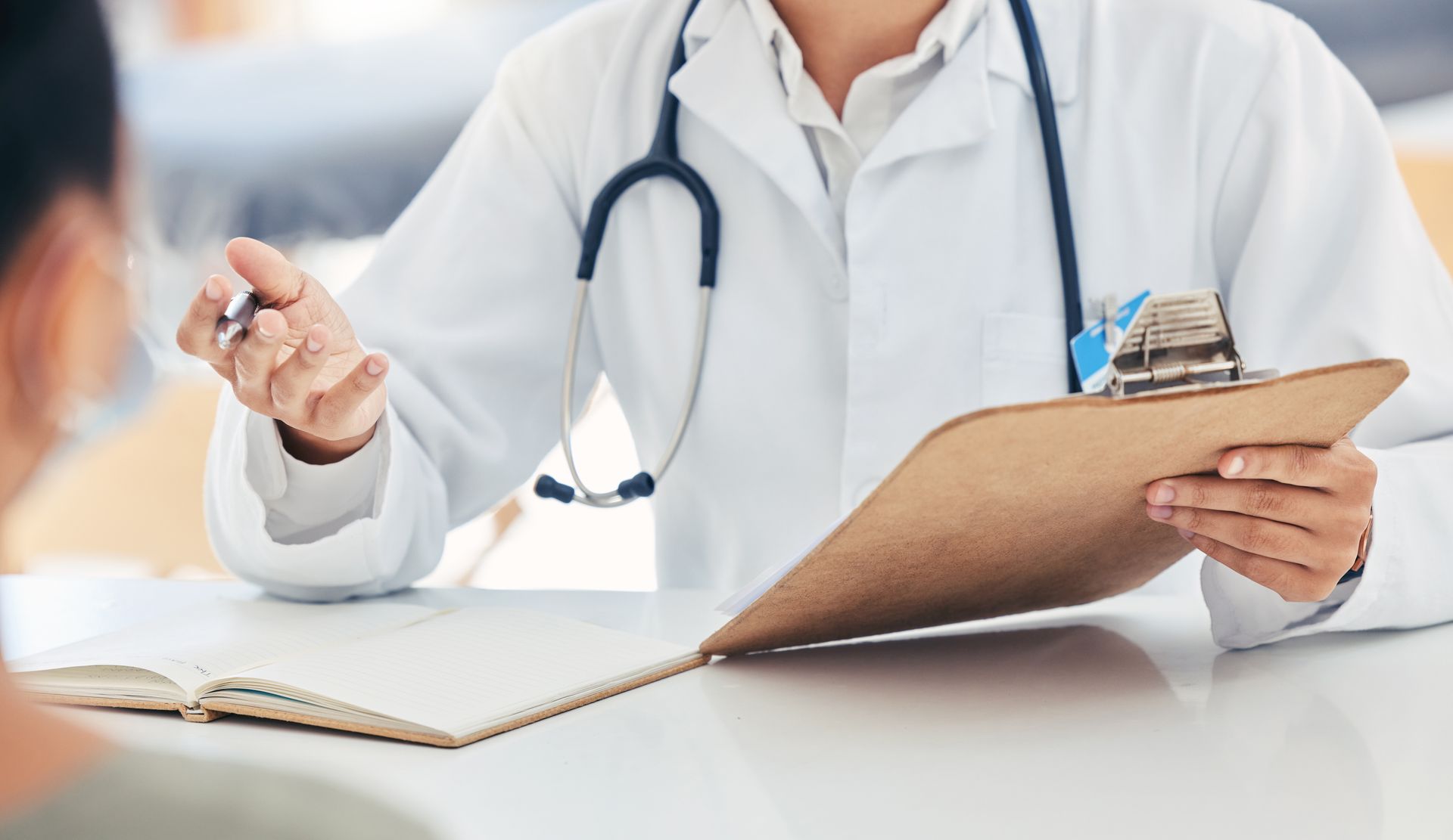 Doctor in white coat with stethoscope, points at clipboard, speaking with patient.