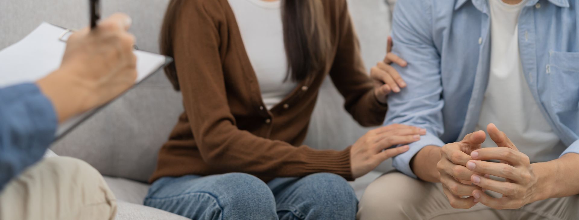 Couple in therapy, one comforting the other with a hand on the arm. Therapist taking notes.