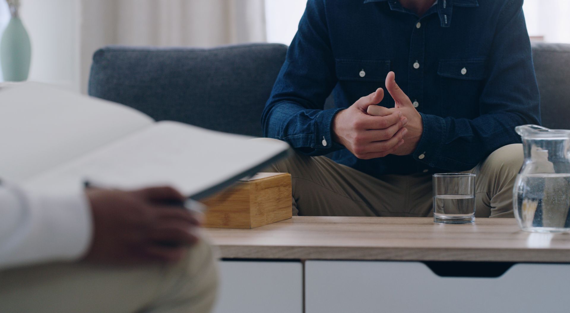 Man in blue shirt, hands clasped, talking to person holding open notebook on a couch.
