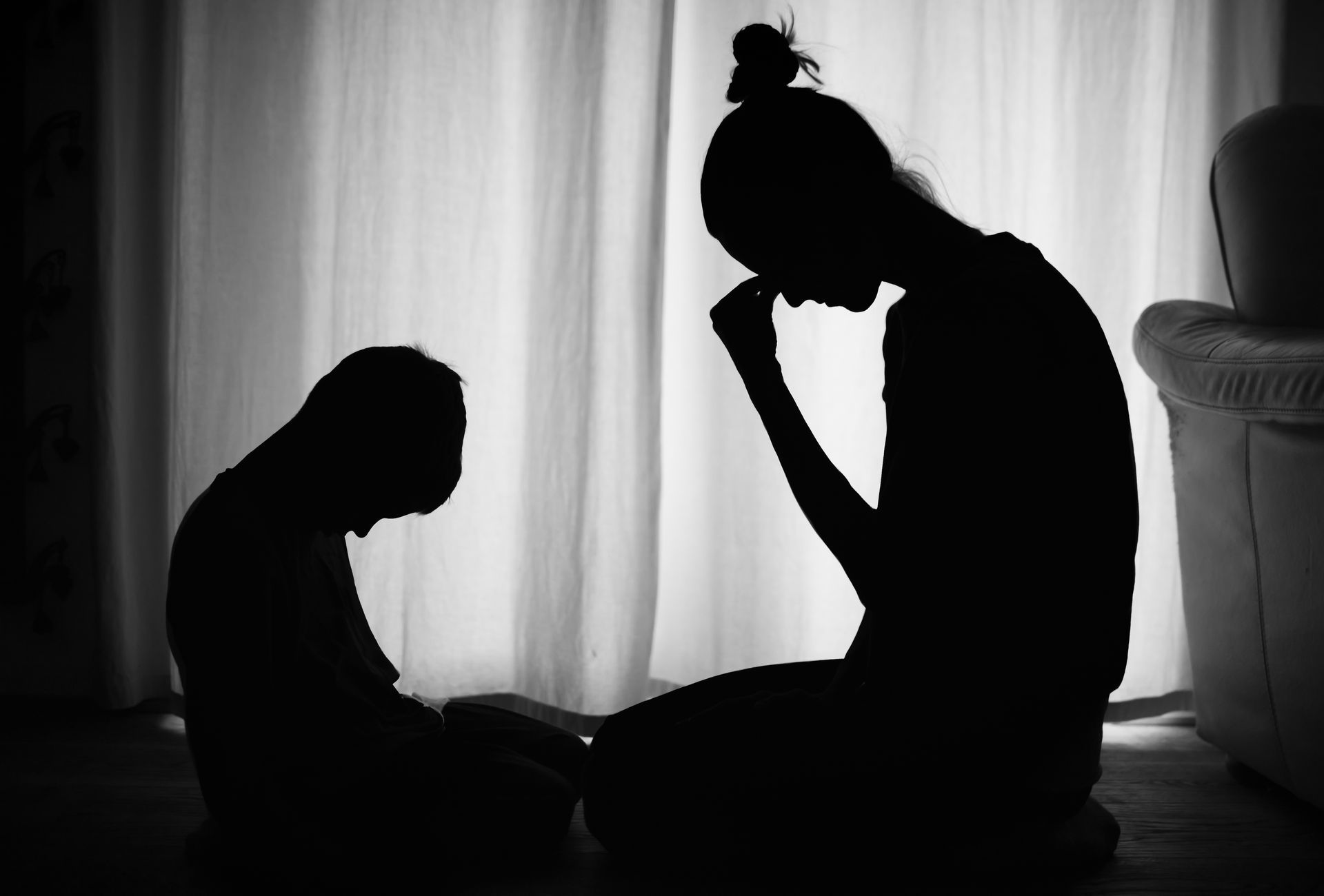 Silhouette of two people, one appearing to be an adult, and one a child, sitting and bowed down in front of a curtain.