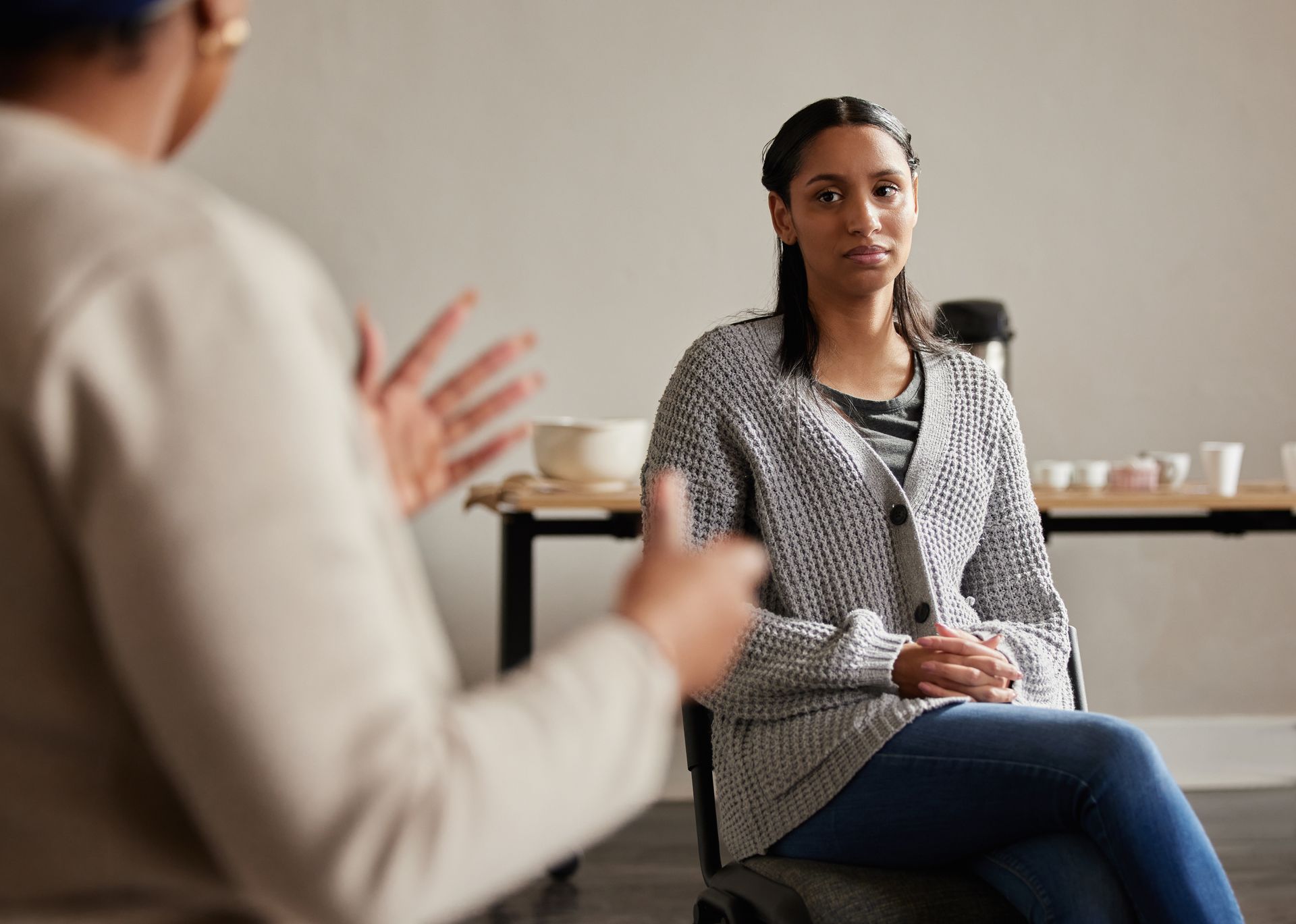 Woman in therapy session, sitting and listening to person speaking, both in a brightly lit room.