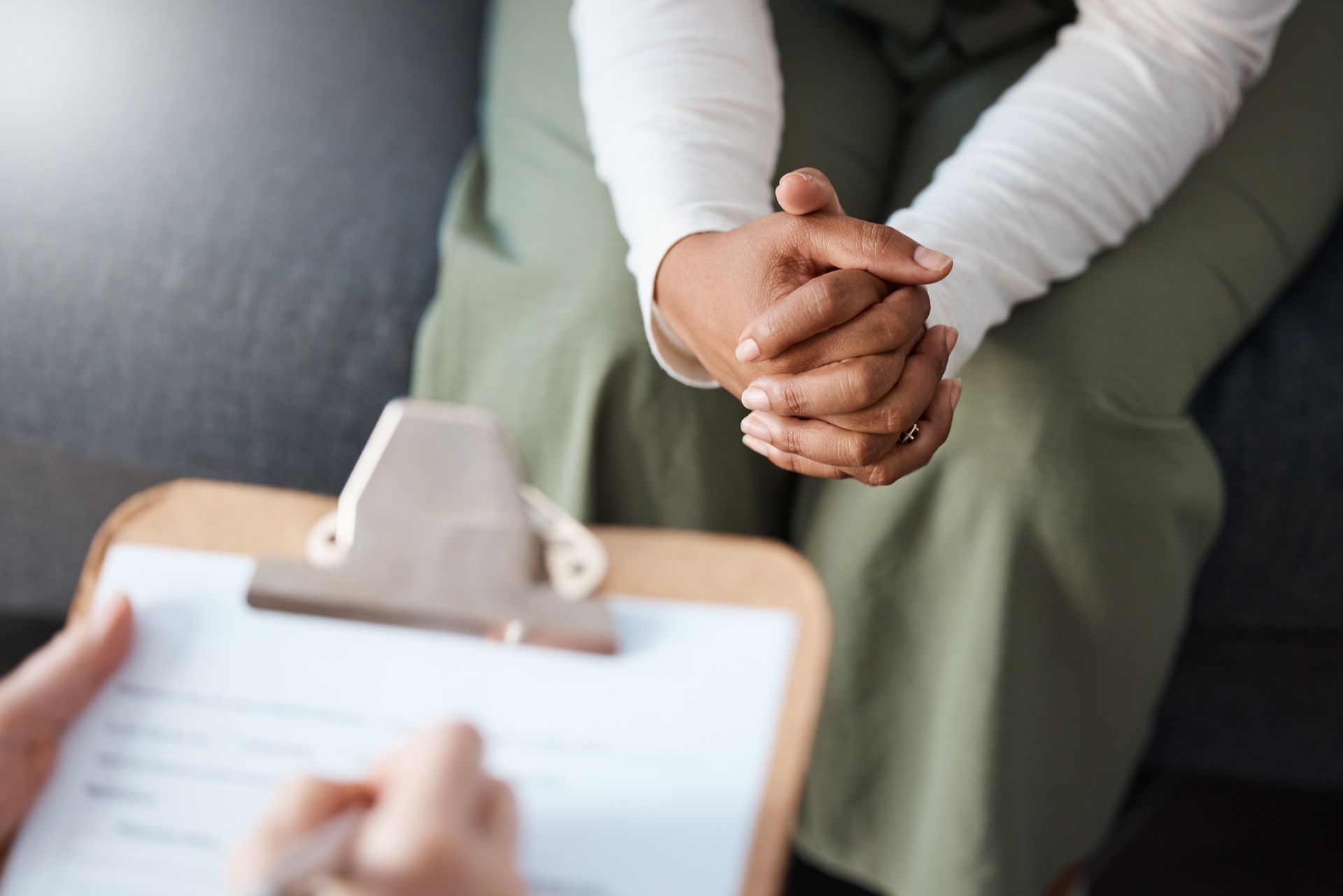Person sitting, hands clasped, facing person writing on clipboard.