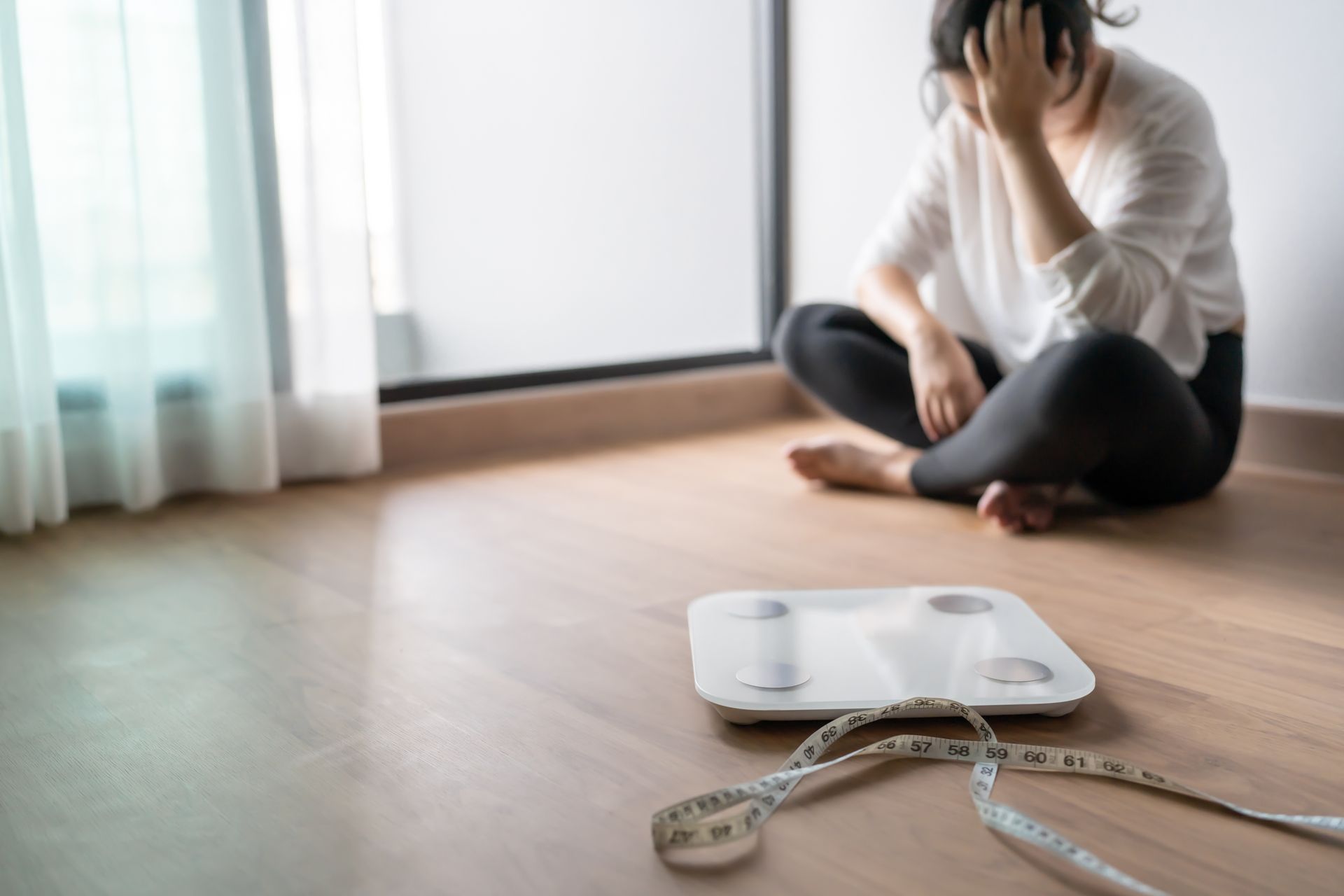 Woman sits on floor, hand on head, near a scale and measuring tape.
