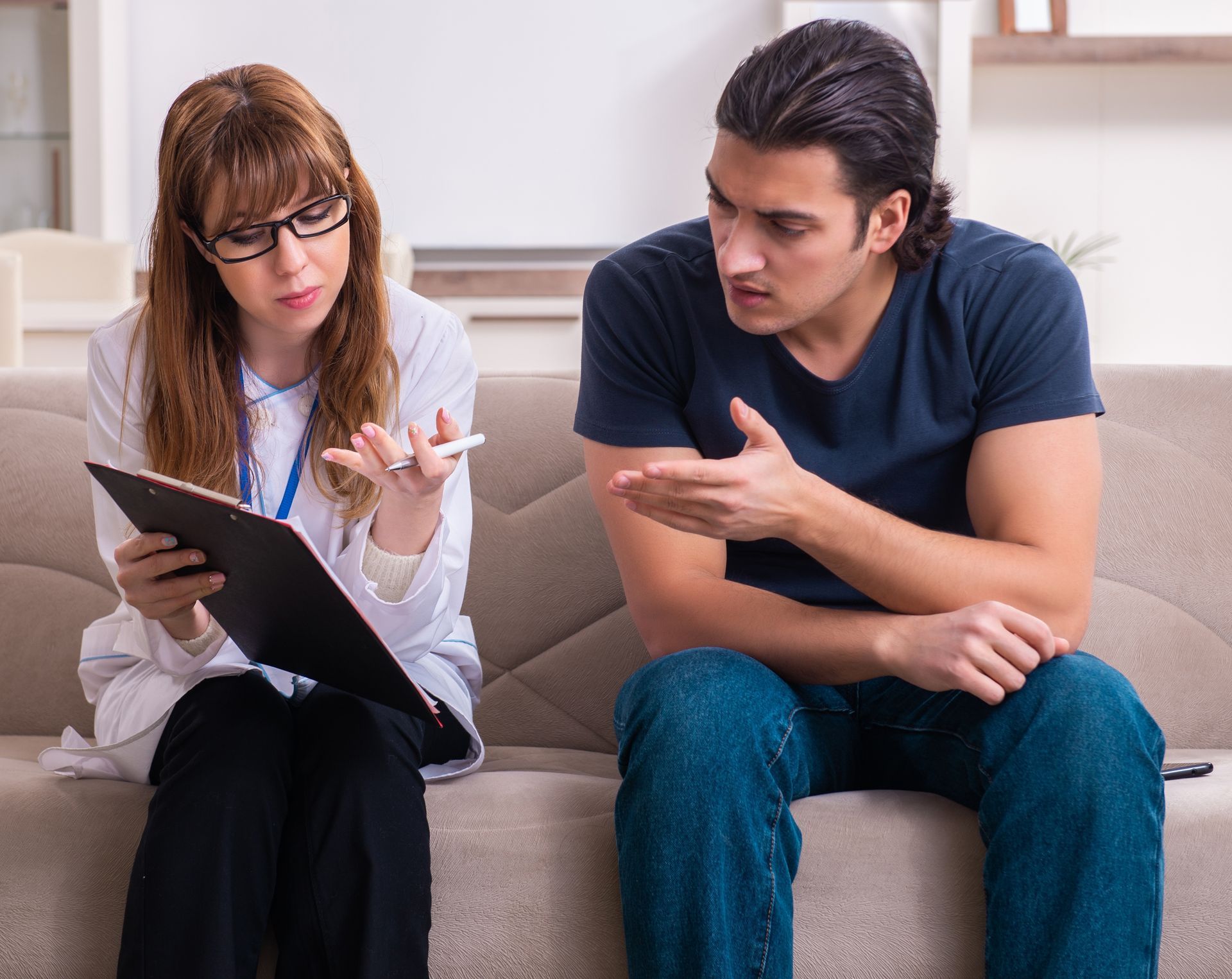 Therapist with glasses takes notes as a client gestures, sitting on a sofa in an office.