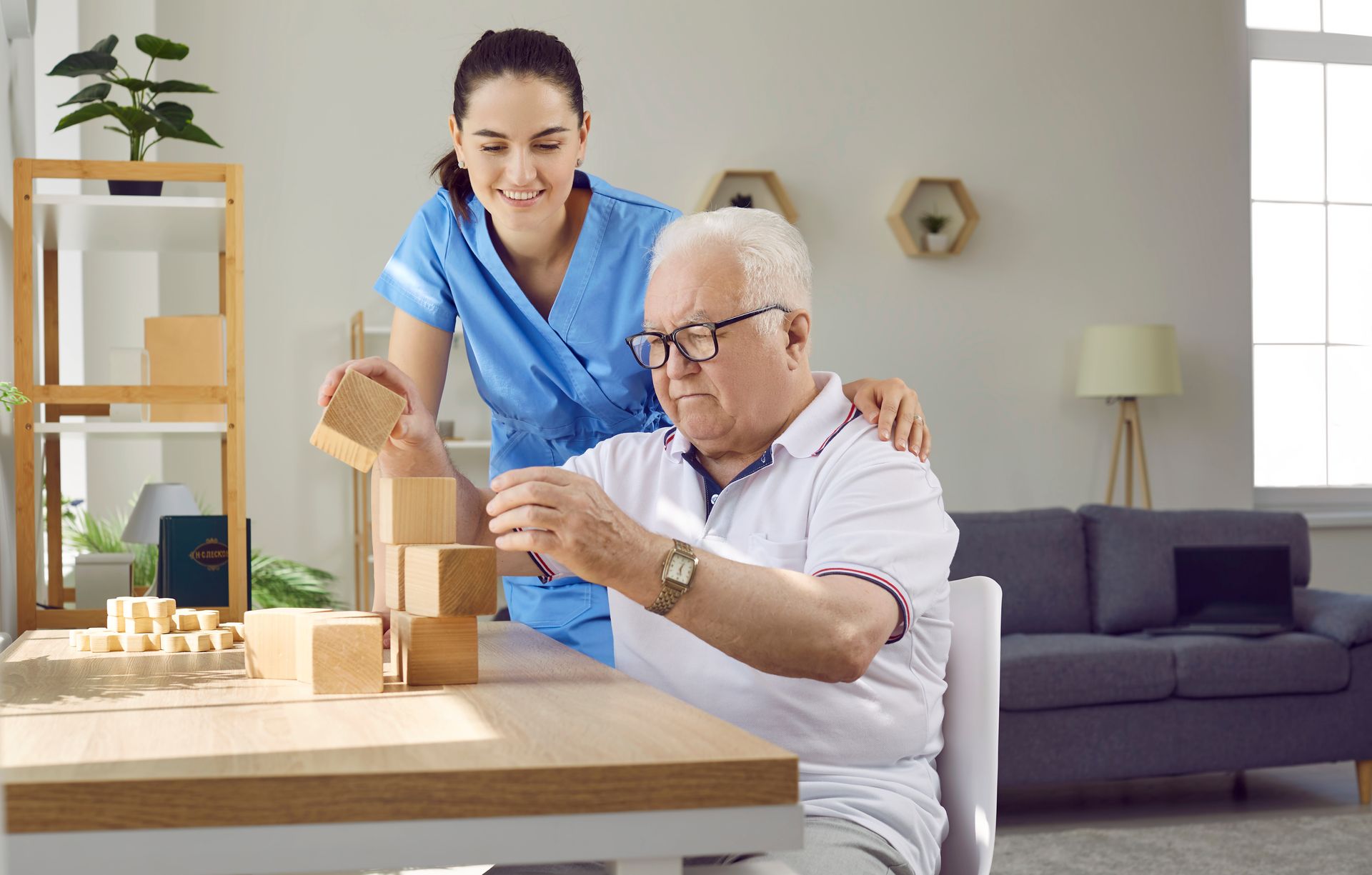 Woman in blue uniform assisting a man with glasses to stack wooden blocks at a table.