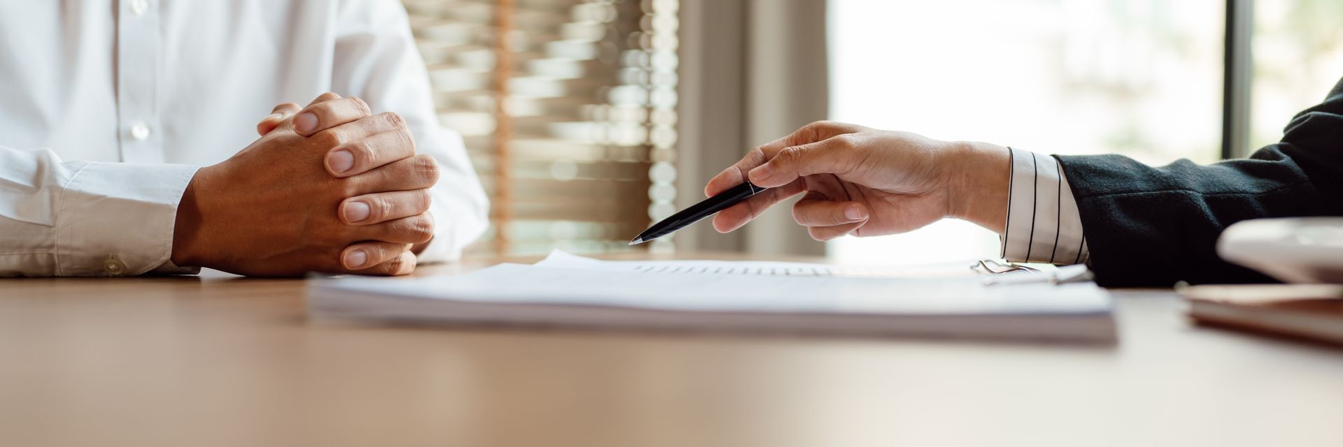 Two people at a table; one points to a document with a pen. The other person’s hands are clasped. Two people at a table; one points to a document with a pen. The other person’s hands are clasped.