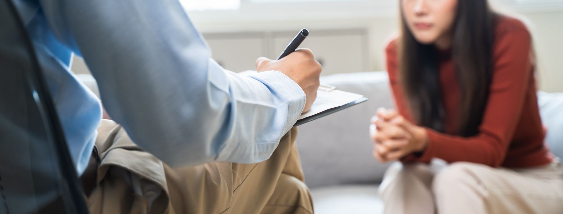 A therapist takes notes while a patient sits on a couch with hands clasped. A person taking notes while a woman sits on a couch, hands clasped.