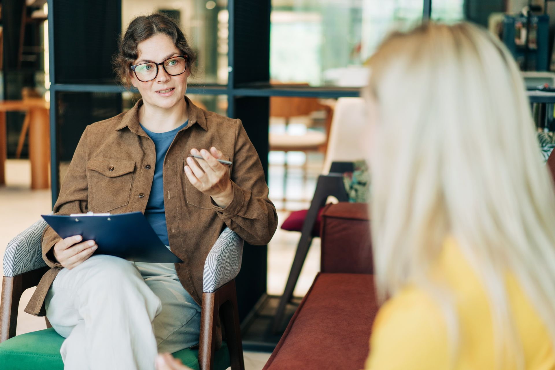 Therapist in glasses, brown jacket, blue shirt, and light pants talks to a blonde client in a bright setting.
