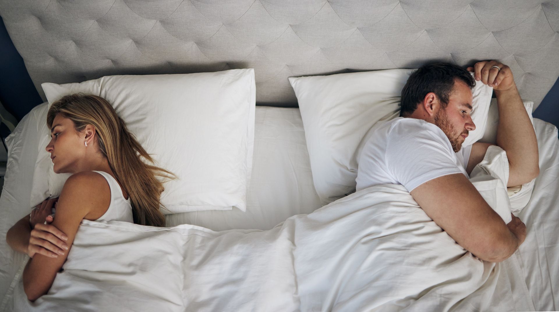 Couple in bed, facing away from each other. White sheets, pillows.