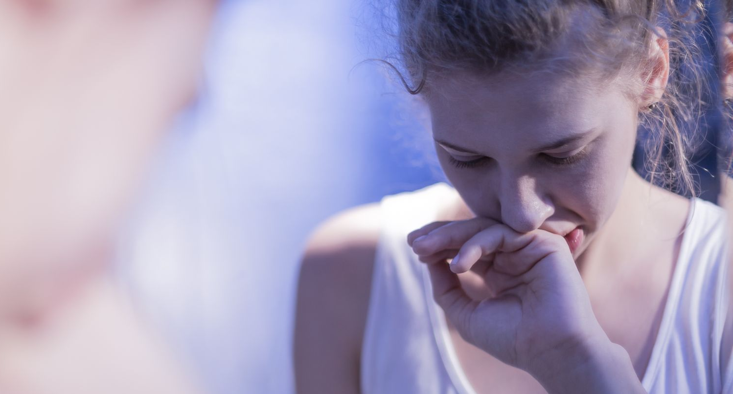 Woman in white top, with hand over mouth, looking down with a sad expression; blurred background.