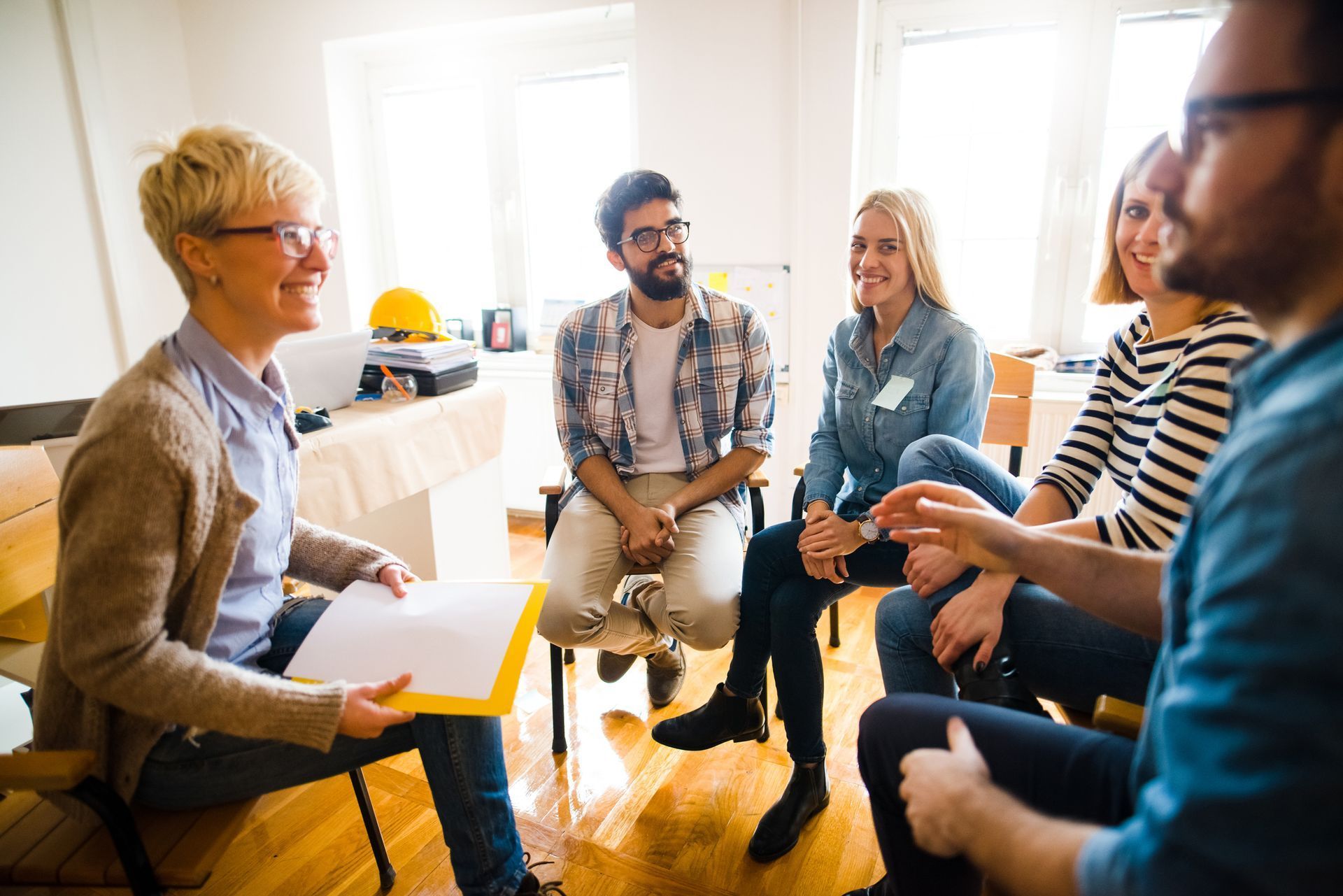 Group therapy session: People seated in a circle, engaged in conversation. A therapist holds notes, smiling.