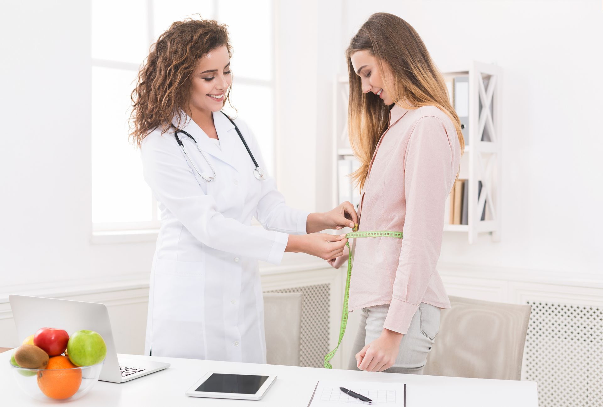 Doctor measuring patient's waist in a bright office. Fruits on desk.