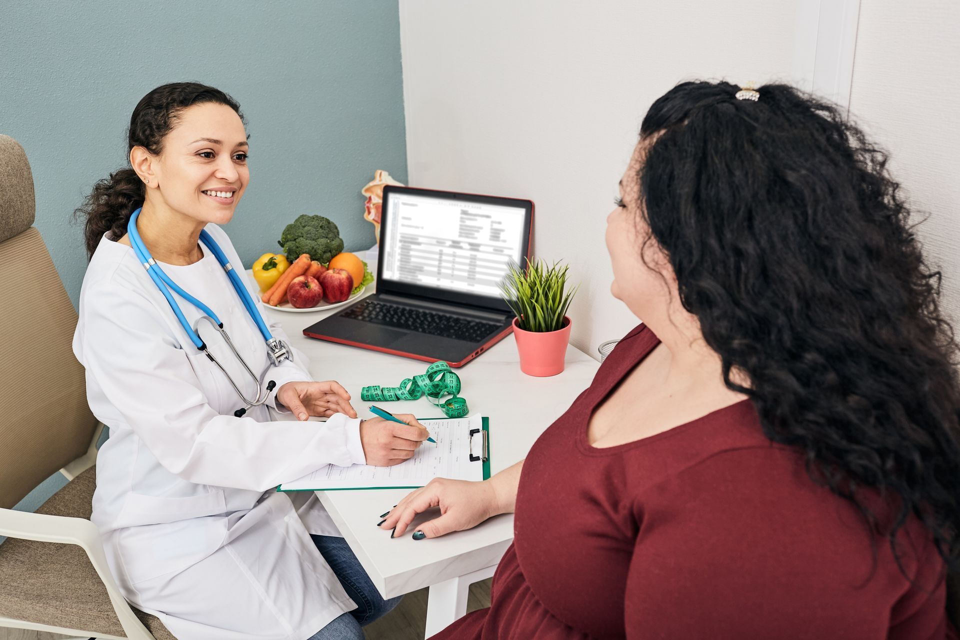 Doctor smiling at patient; examining notes. Laptop and fruits on desk.