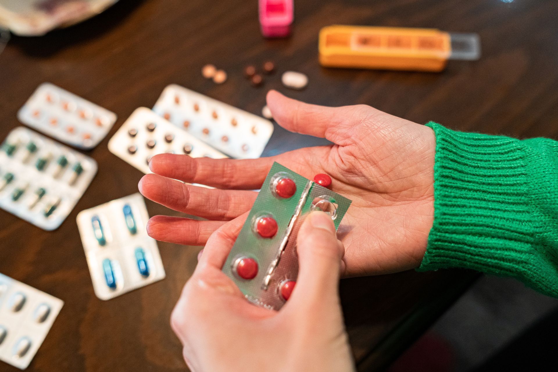 Person holding red pills in blister pack, surrounded by other medications on a table.
